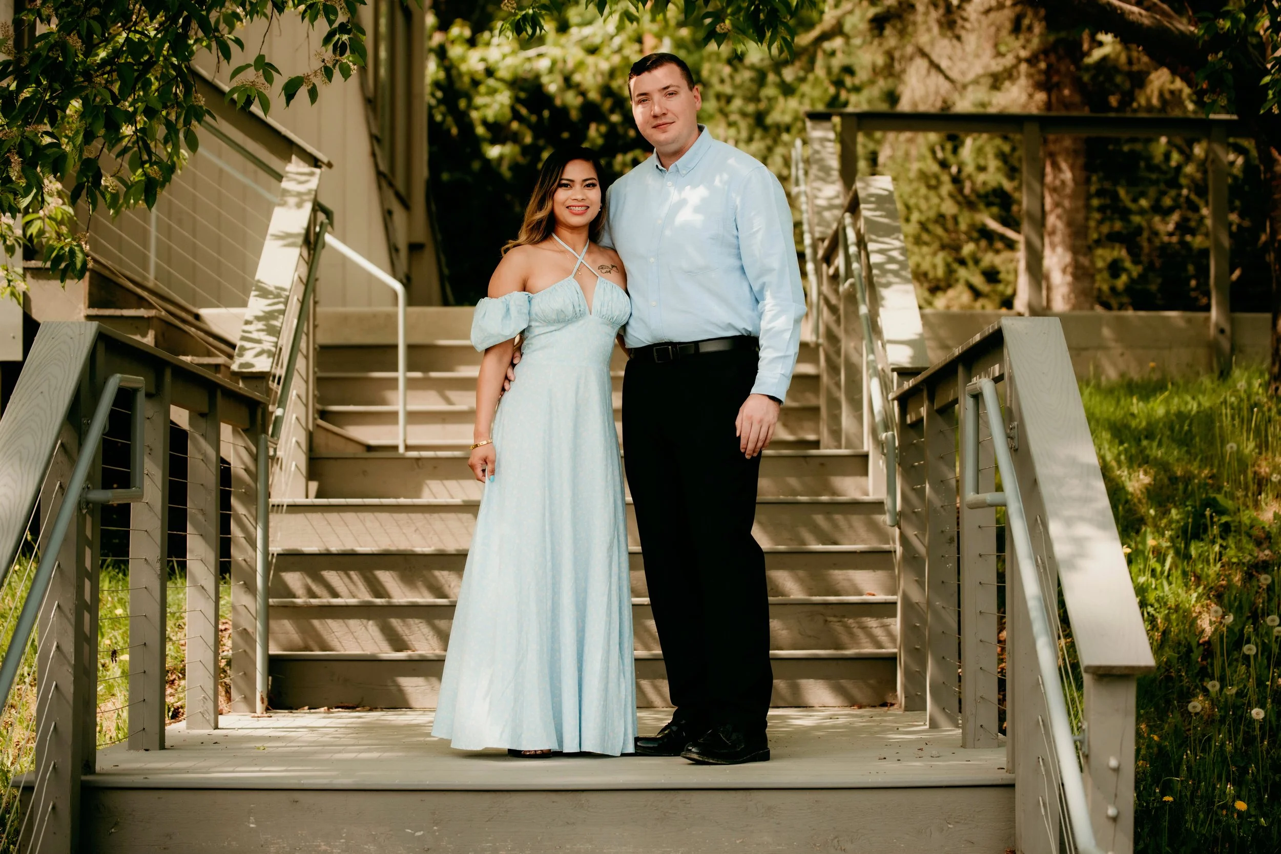 A couple standing on a wooden staircase outdoors, with trees and greenery in the background. The woman is in a light blue off-the-shoulder dress and the man in a light blue shirt and black pants, both smiling.