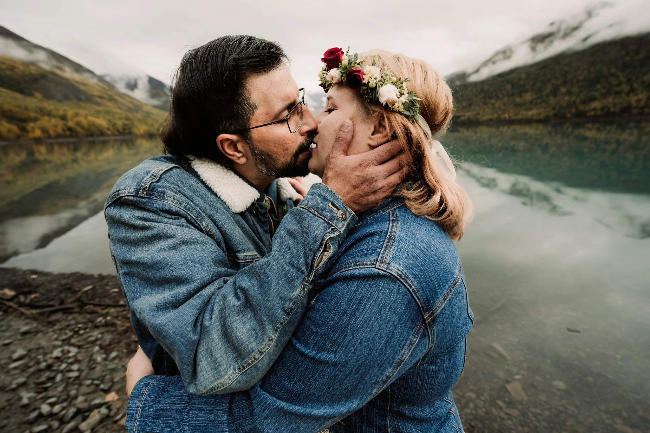 A couple sharing a kiss by a scenic lake with mountains in the background, the woman wearing a flower crown.