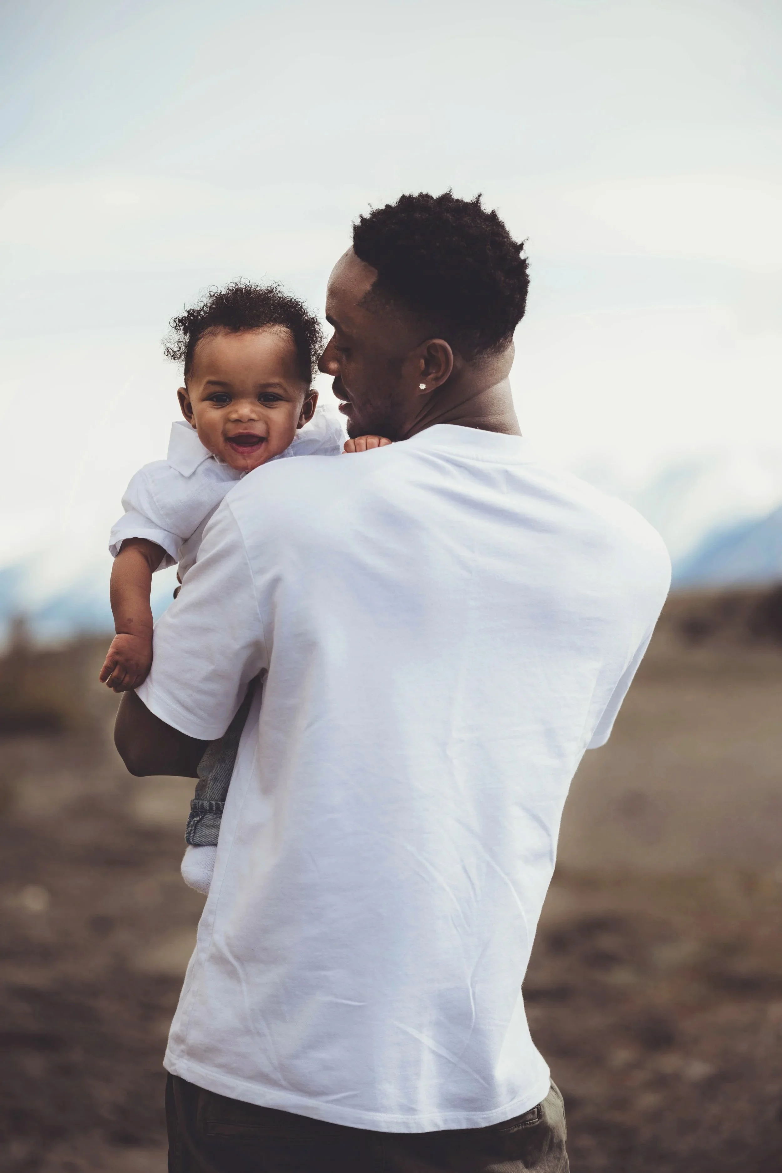 A man holding a smiling baby outdoors with a cloudy sky in the background.