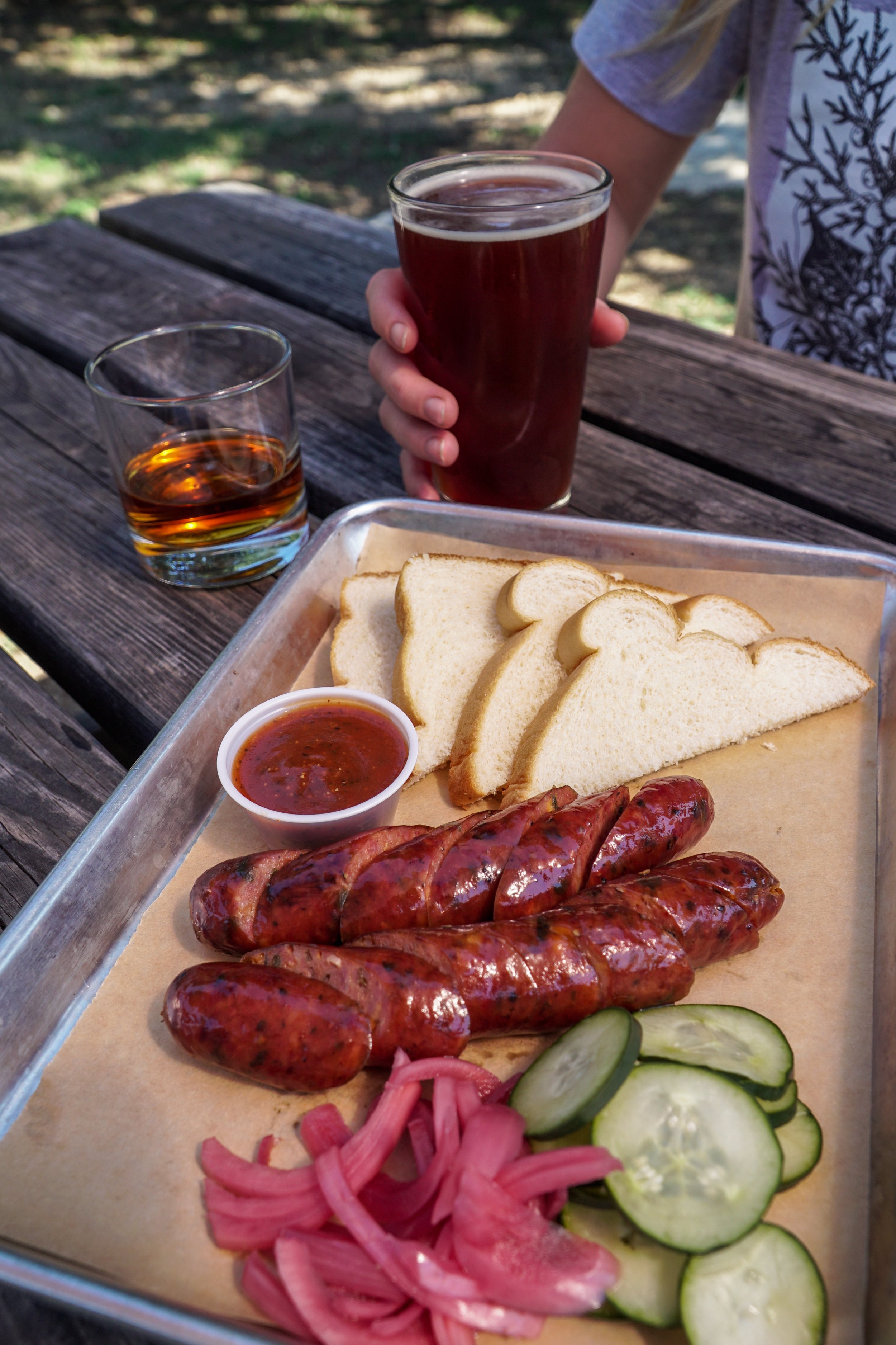 plate of sliced sausage with side of bread, pickles, pint of beer and glass of Treaty Oak whiskey