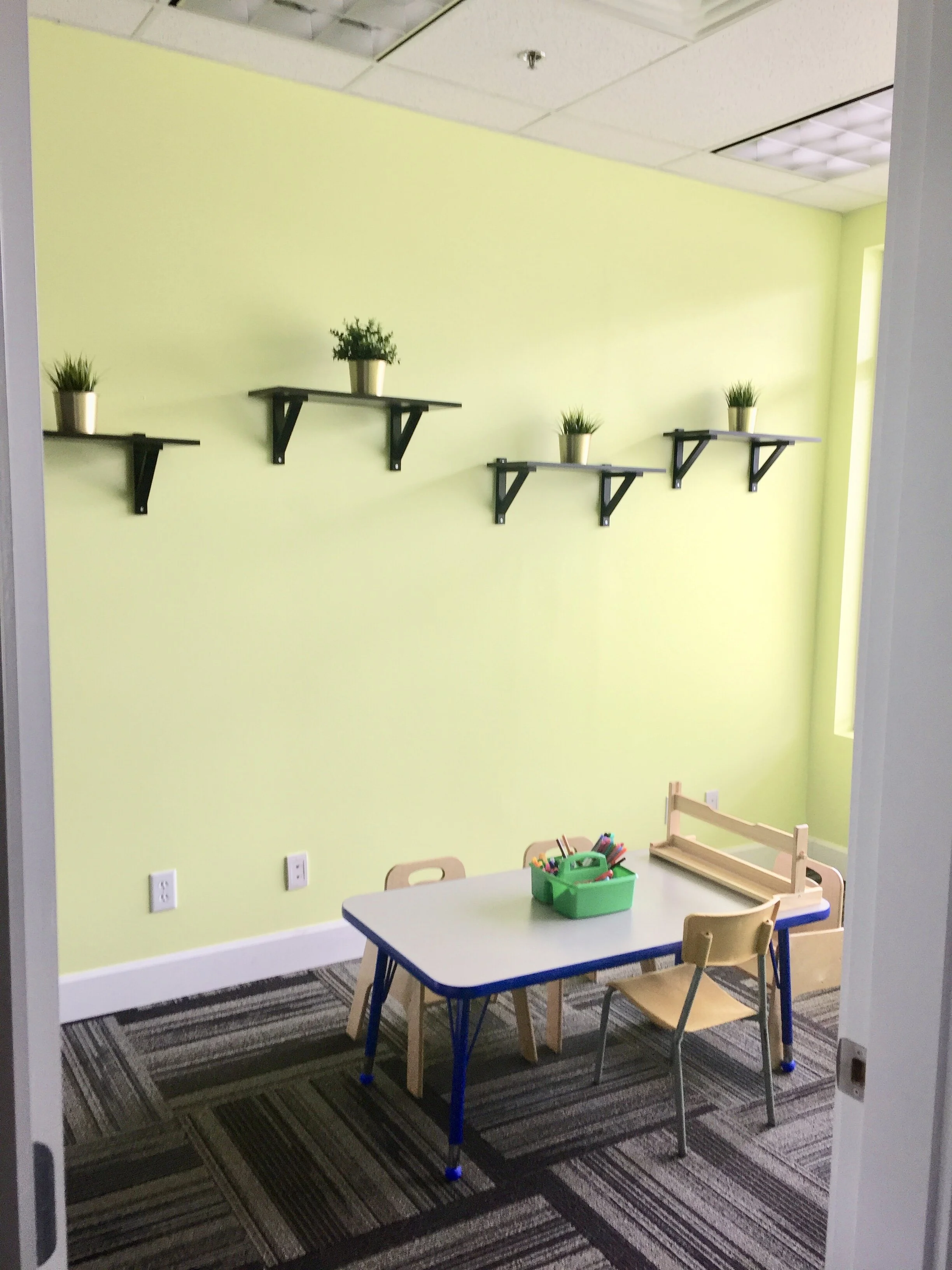Small playroom with light green walls and a carpeted floor. There is a white table with wooden chairs, a green container filled with art supplies, and a wooden toy on the table. Four small black shelves on the wall hold potted plants.