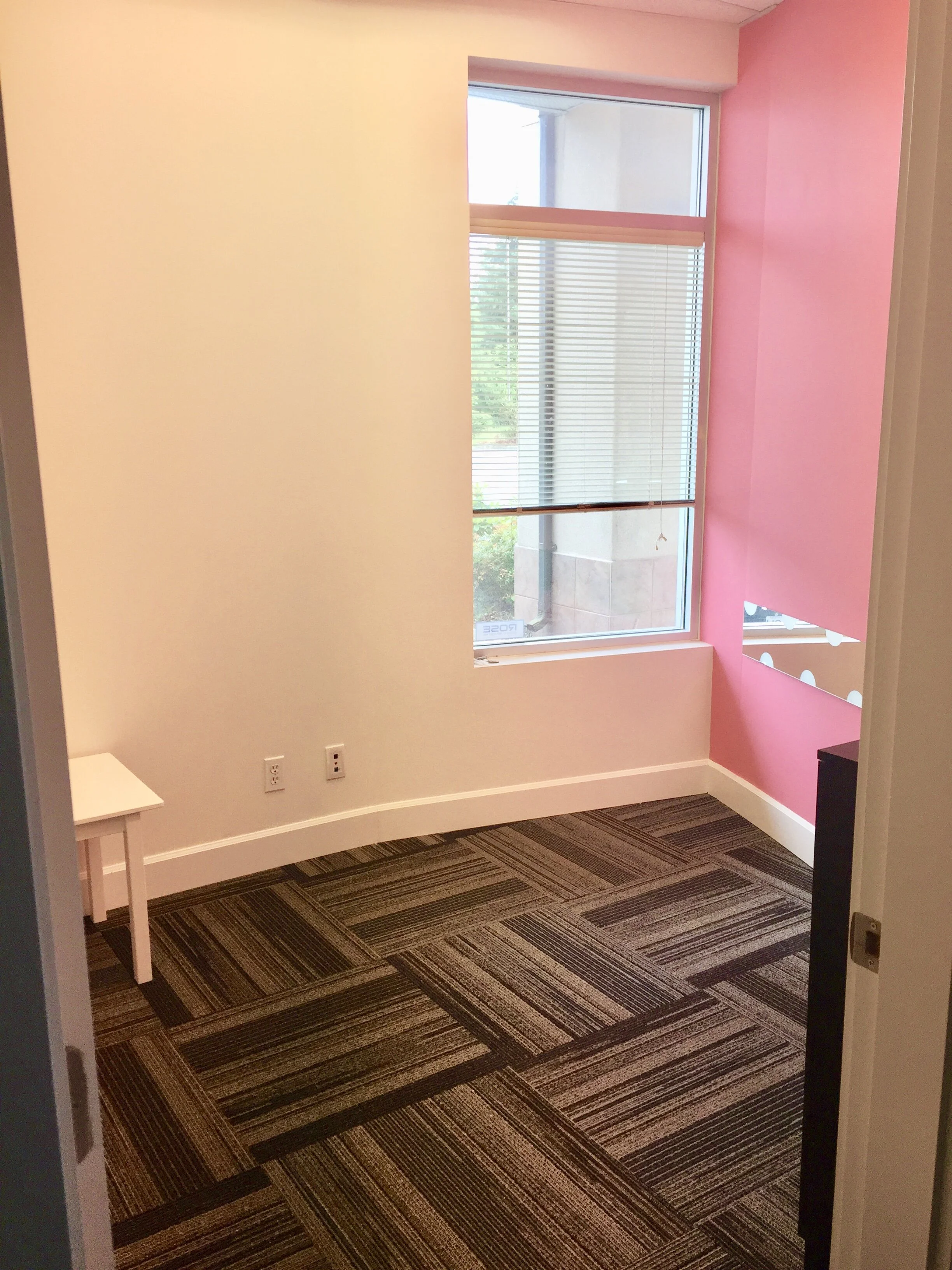 Empty office space with beige and pink walls, a large window with blinds, a patterned carpet, and a small white table in the left corner.