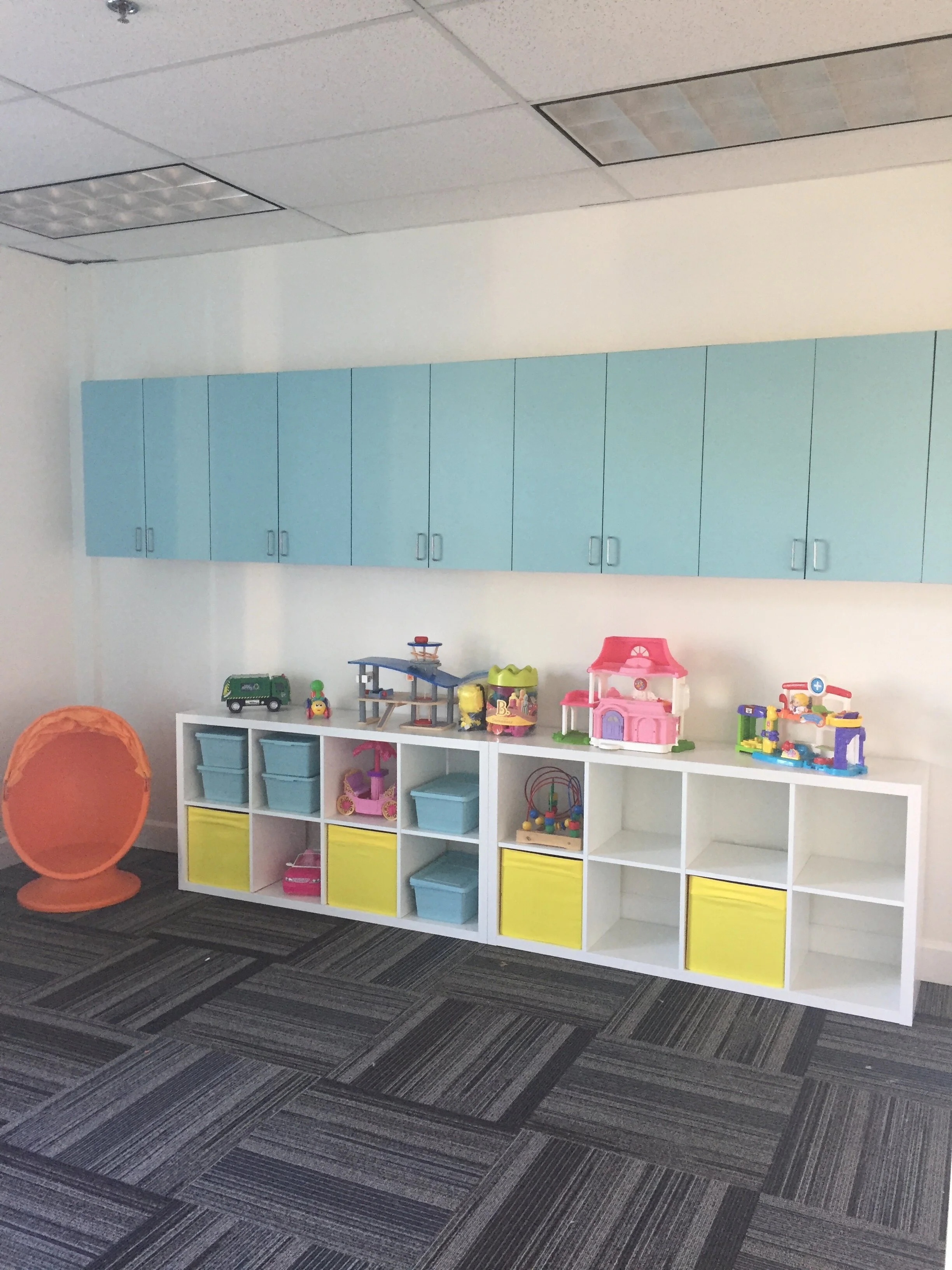 A children's playroom with blue wall cabinets, a white storage unit with colorful bins and toys, including a pink dollhouse, a ride-on toy, and various miniature vehicles, on a patterned carpet floor.