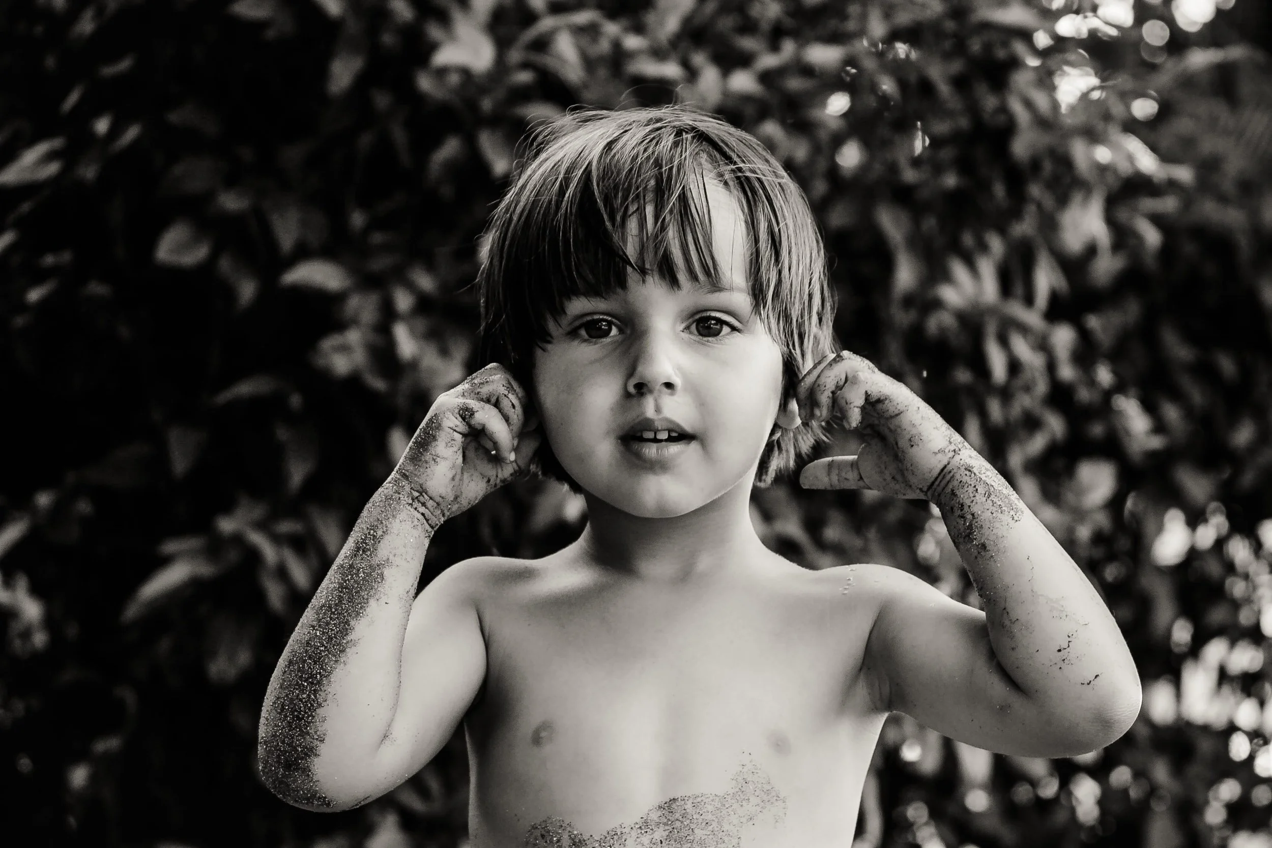 A black and white photo of a young boy with dirty hands and arms, covering his ears with his fingers. He has short, messy hair and is looking directly at the camera with a neutral expression, against a background of blurred foliage.
