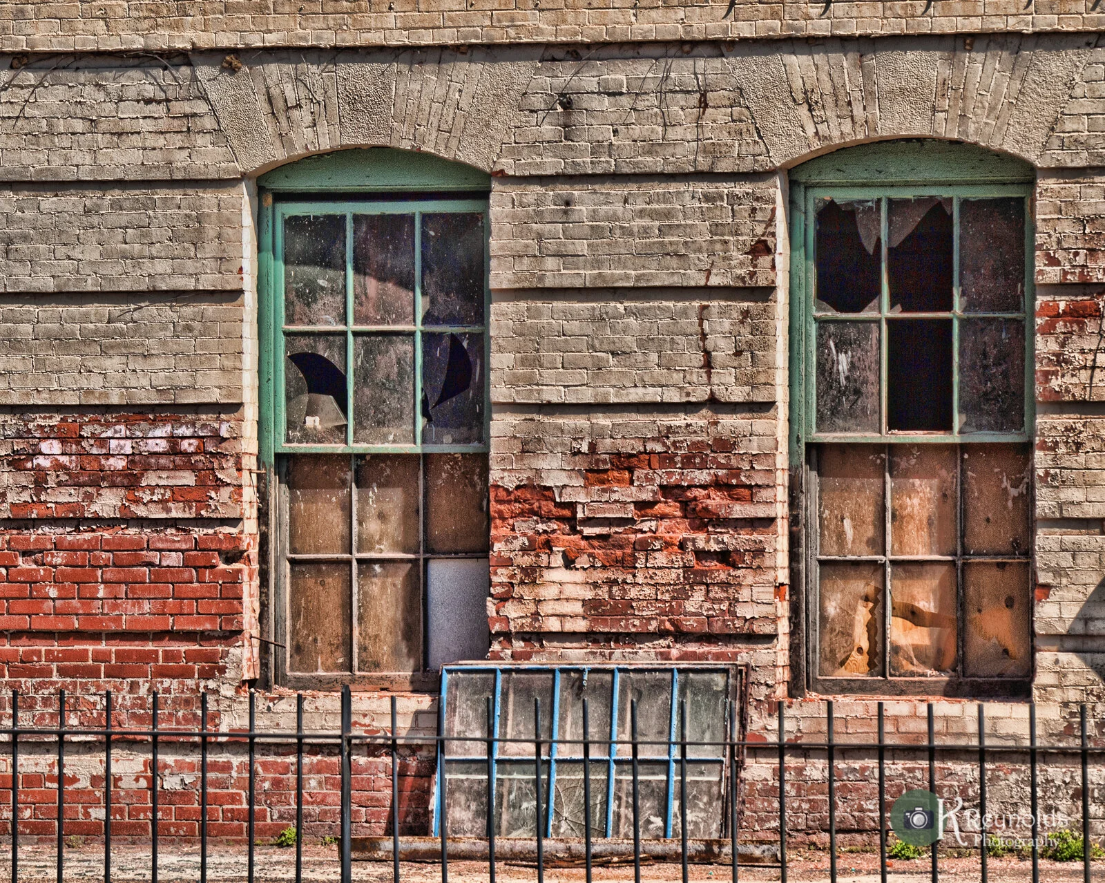 An old brick building with two broken windows and a black metal fence in front. The bricks are weathered, with some portions painted red and peeling paint near the windows.