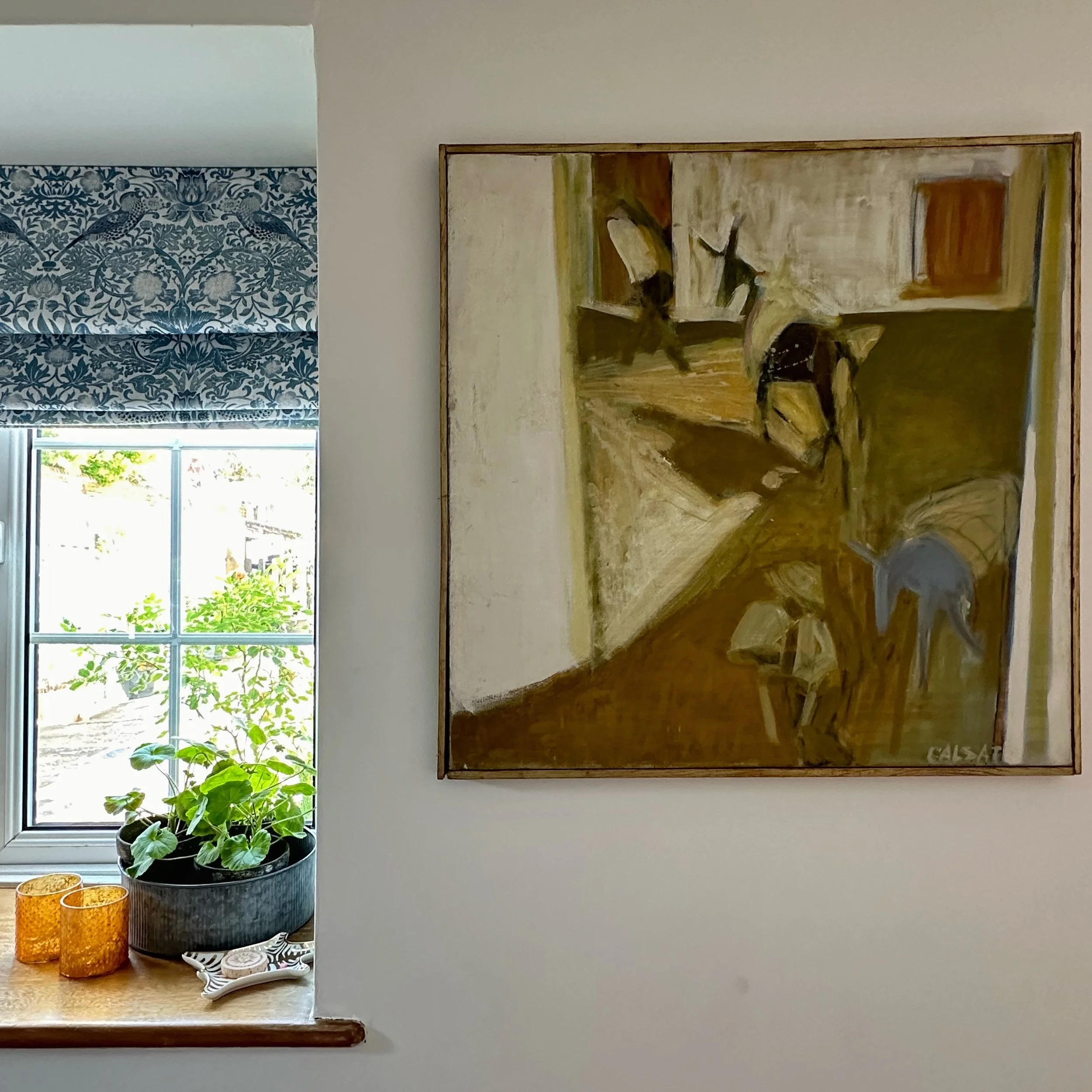 Framed artwork displayed beside a cottage window with patterned Roman blind and houseplants.