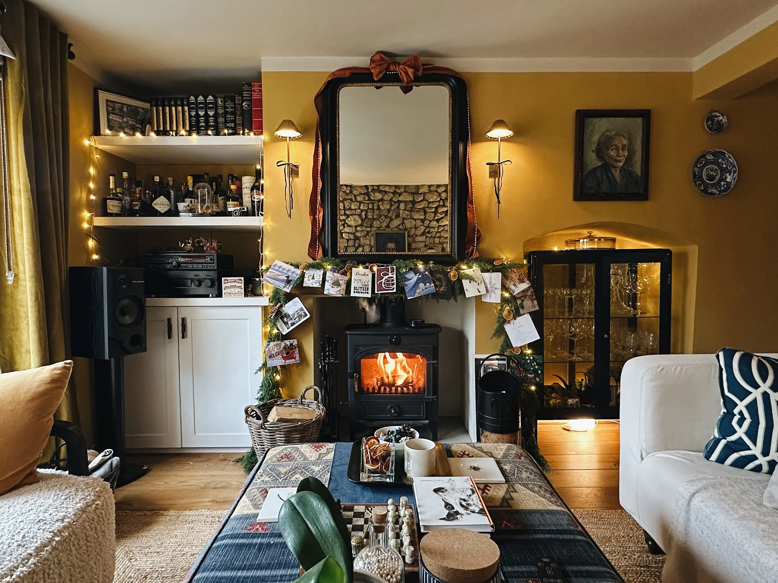 Cottage living room at Christmas with wood-burning stove, built-in shelving, layered textiles and collected artwork.