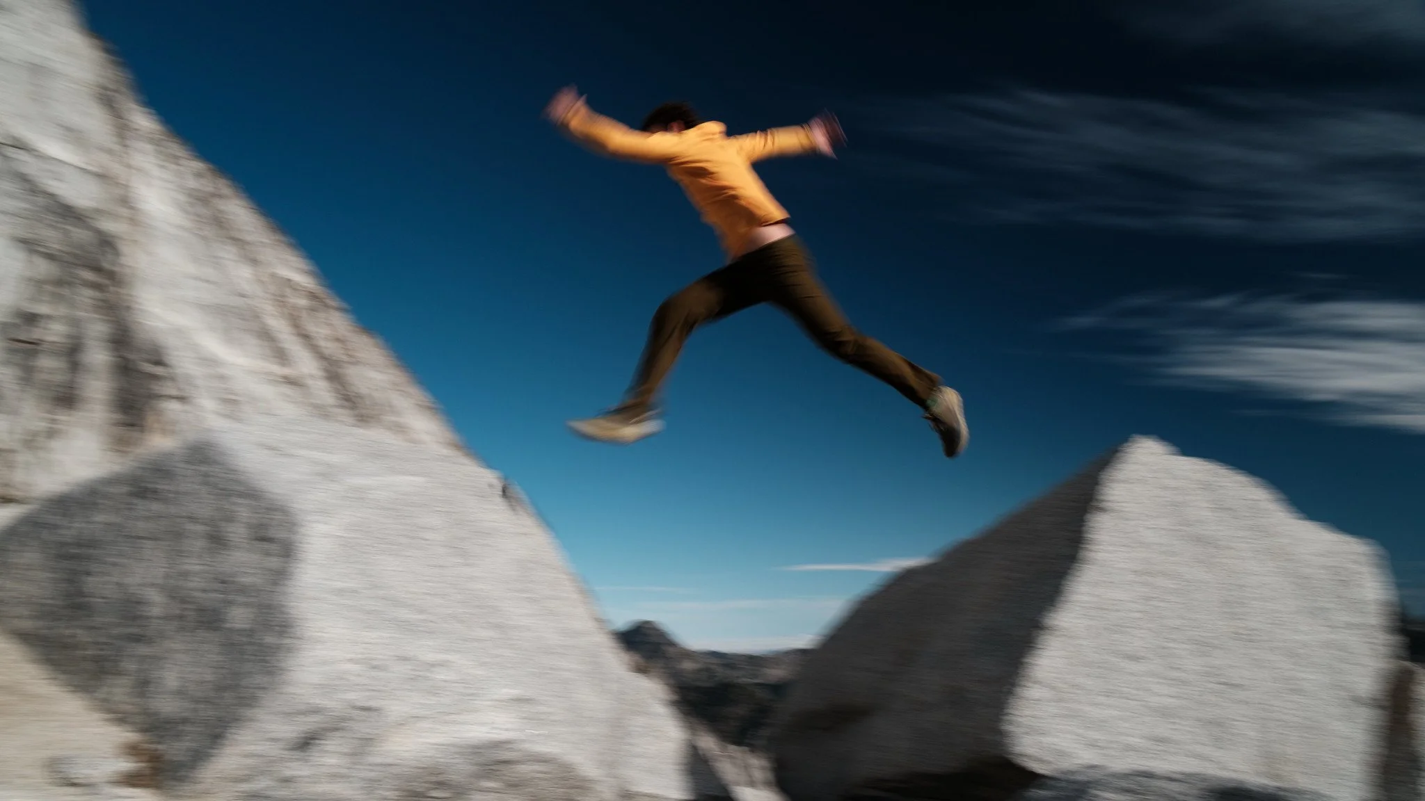 Person jumping between large white rocks outdoors with a blue sky and wispy clouds above.