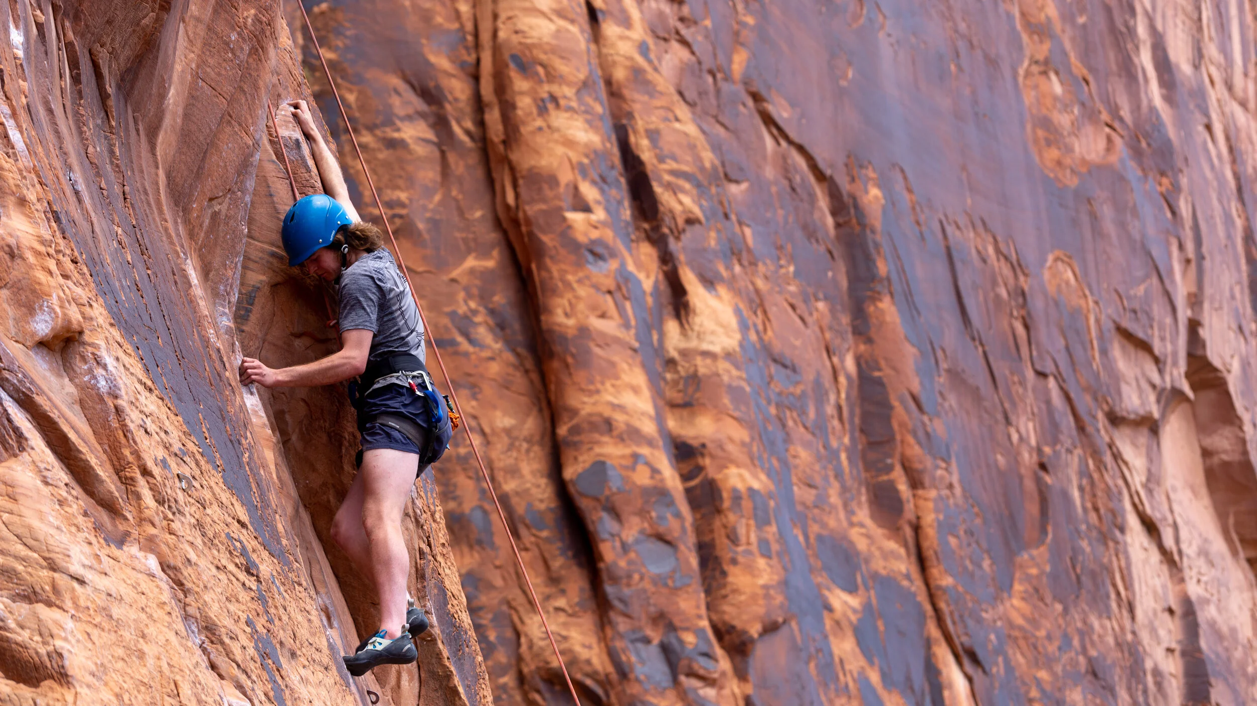 A person rock climbing on an orange sandstone cliff wearing a blue helmet and harness in Moab Utah