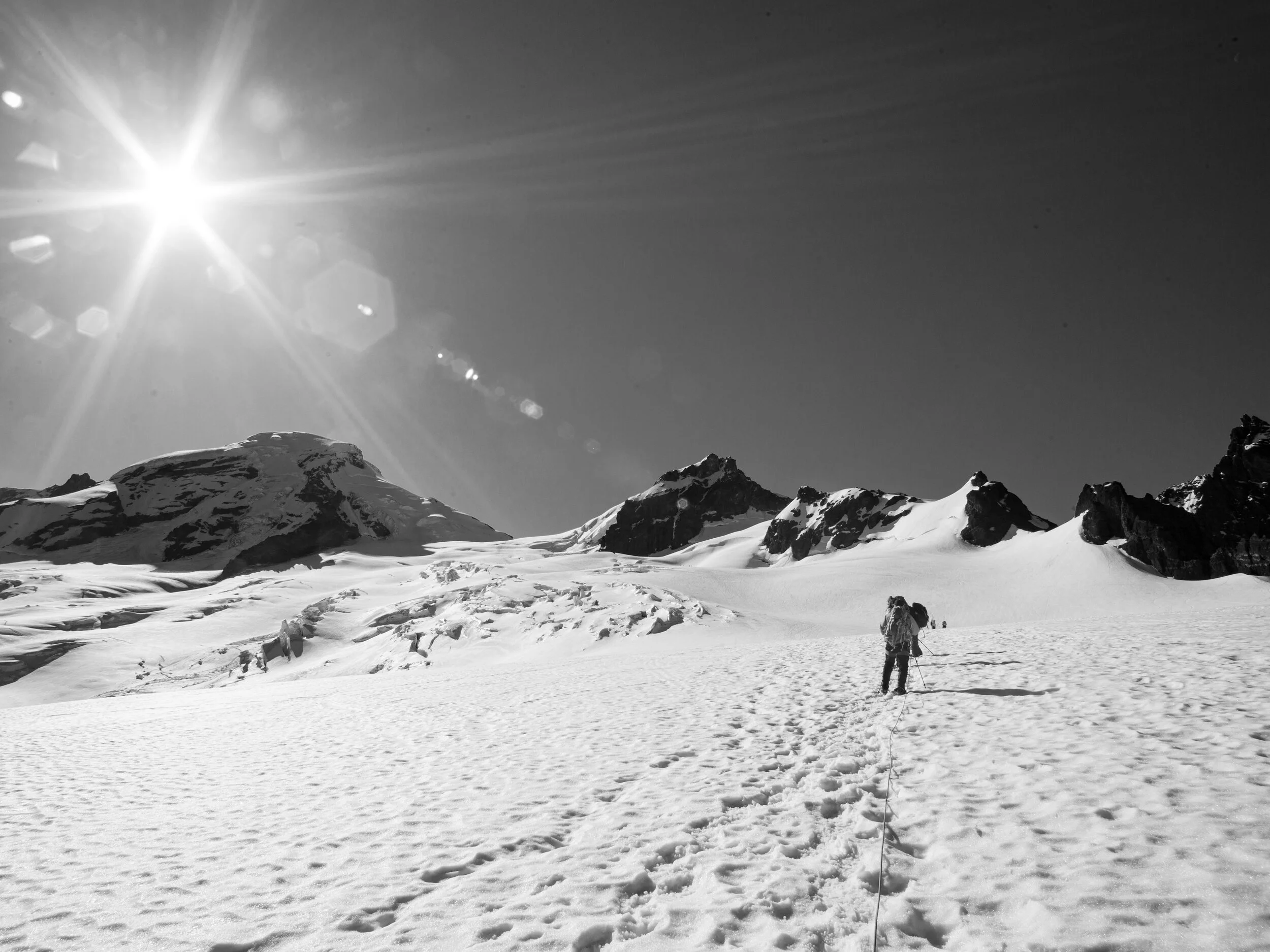 A person hiking on Mount Baker, as they make their way across a glacier snowy mountain terrain under the bright sun.
