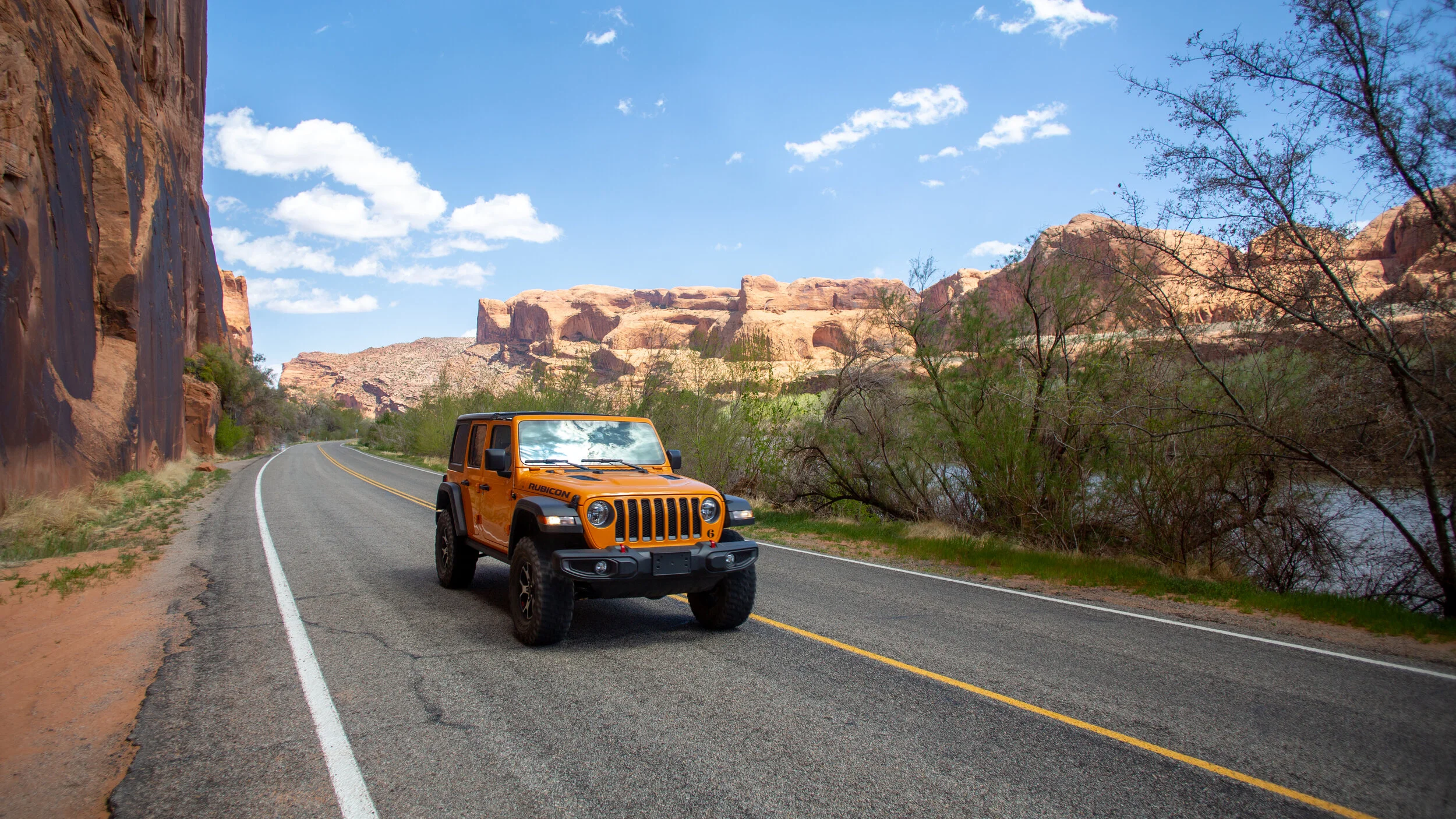 An orange Jeep Rubicon driving on a winding road through a desert landscape with red rock cliffs, sparse trees, and a river in the background under a blue sky with white clouds.