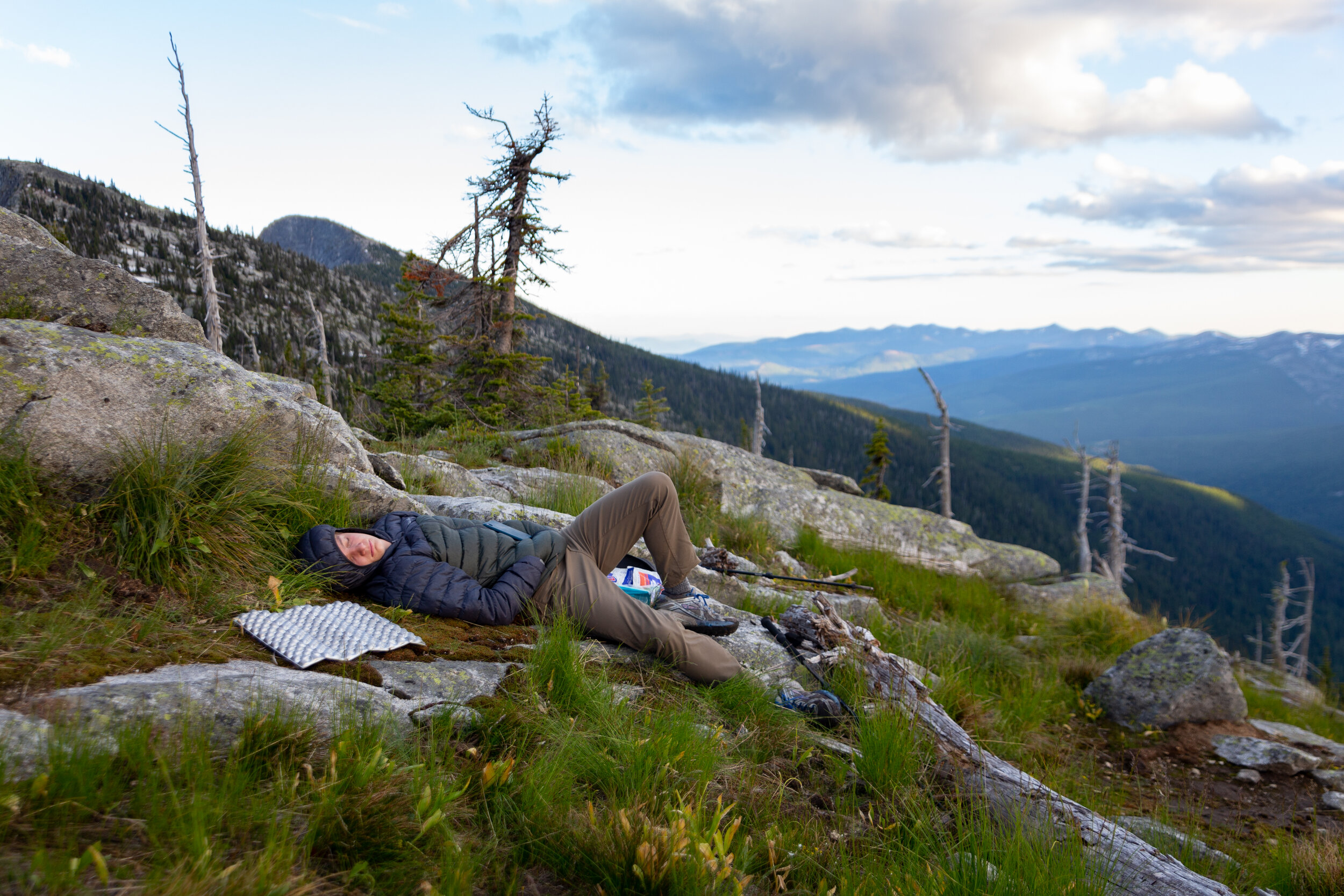 A person sleeping on a grassy and rocky hillside in a mountainous forest during daytime, with cloudy sky and distant mountain range in the background.