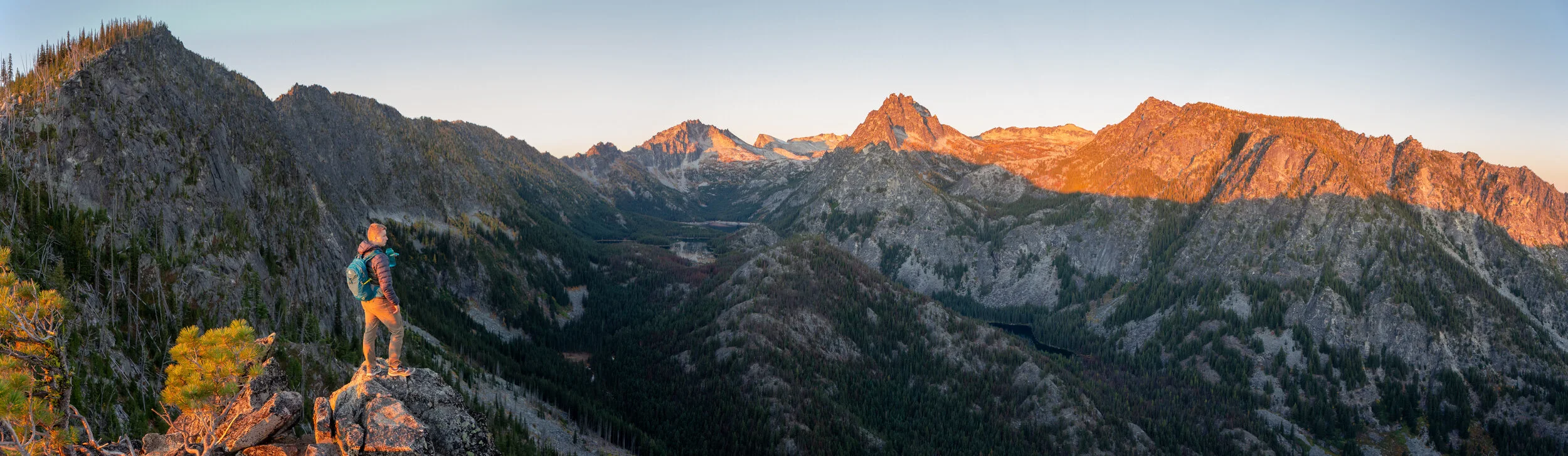 A hiker standing on a rock ledge overlooking a mountain valley at sunset with rugged peaks in the background.