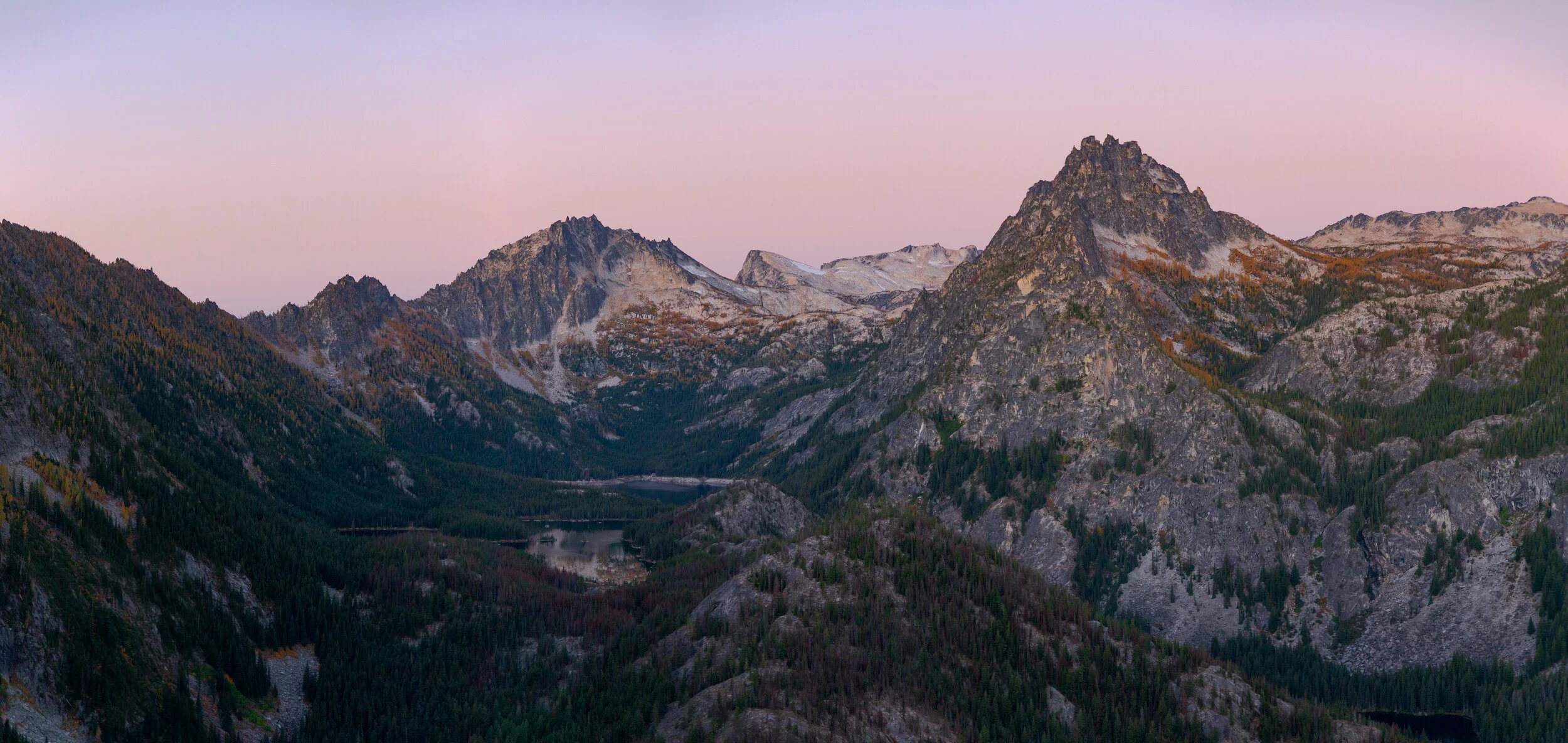 Mountain range with rocky peaks and dense forested slopes at dusk, with a pink and purple sky.