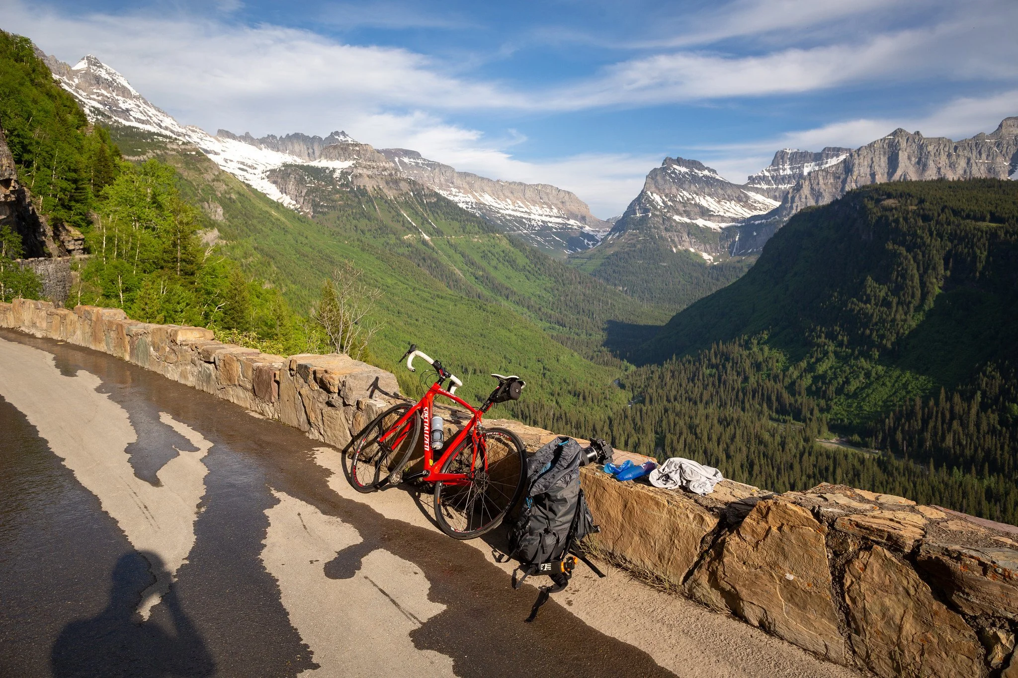 A red bicycle leaning against a stone barrier on a mountain road, with a backpack and clothes nearby, overlooking a lush green valley with snow-capped mountains in the distance under a partly cloudy sky.