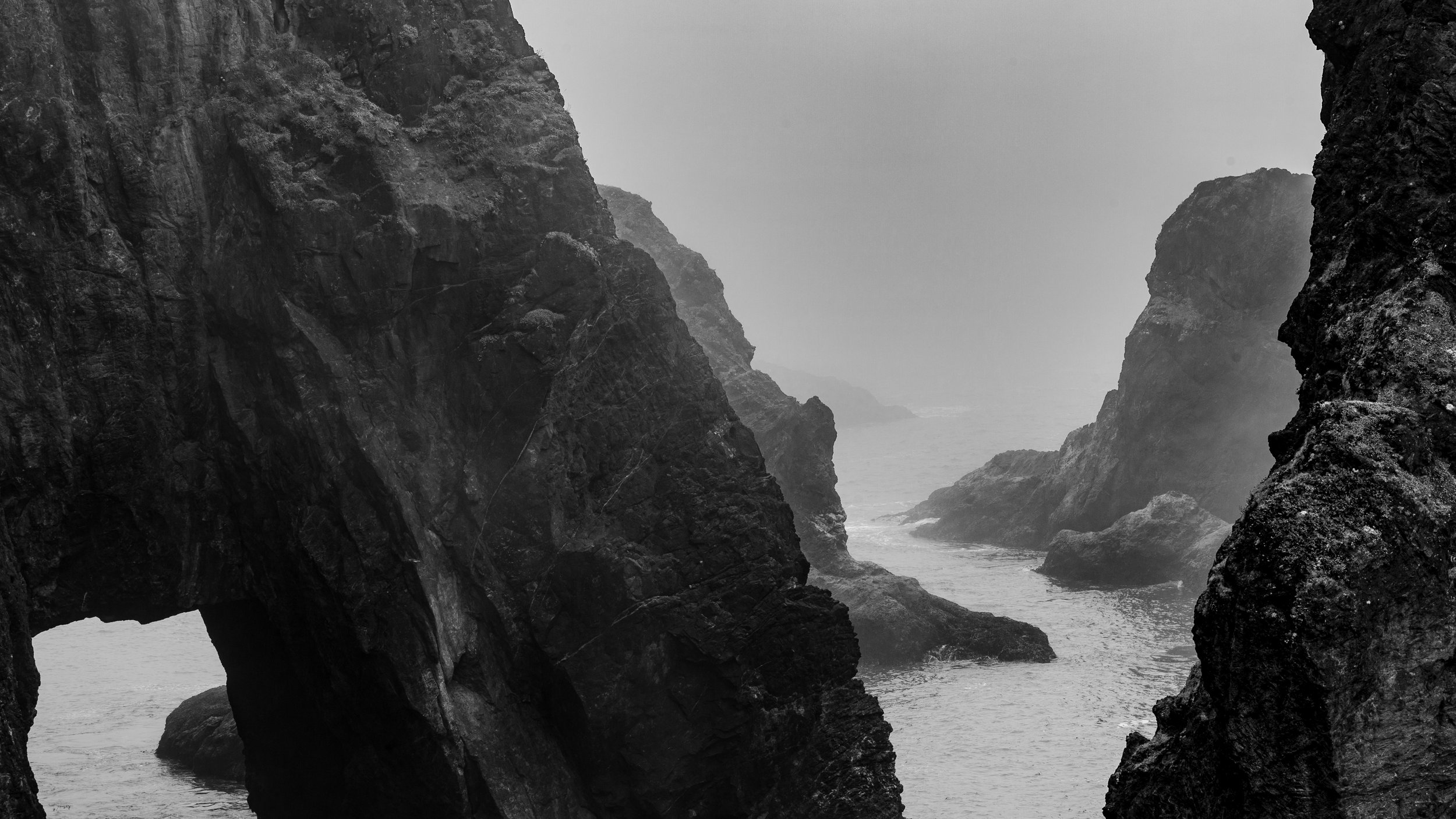 Black and white photograph of rugged coastal cliffs with water between them, shrouded in mist.