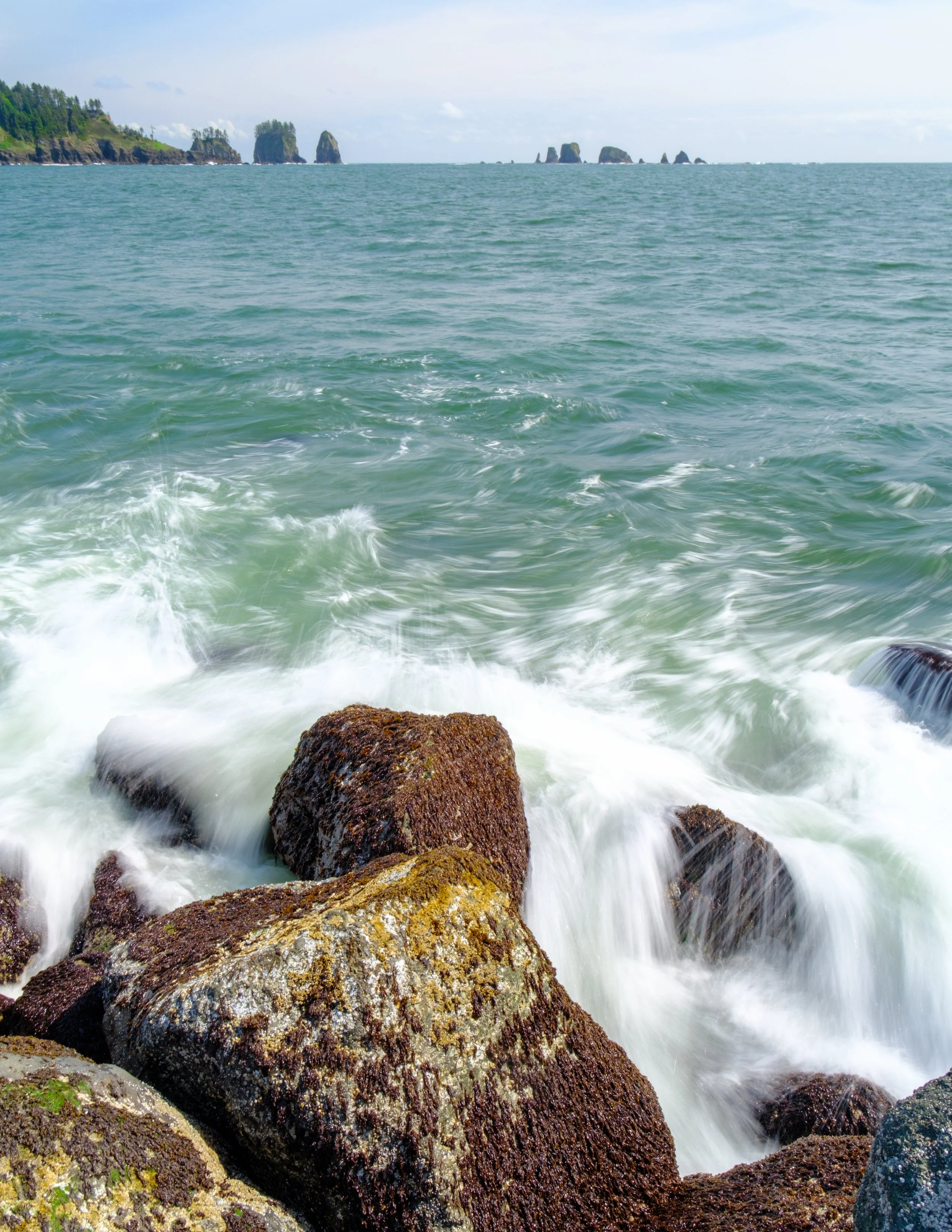 Ocean waves crashing against rocks near the shoreline with distant rocky islands and green trees along the coast under a partly cloudy sky.