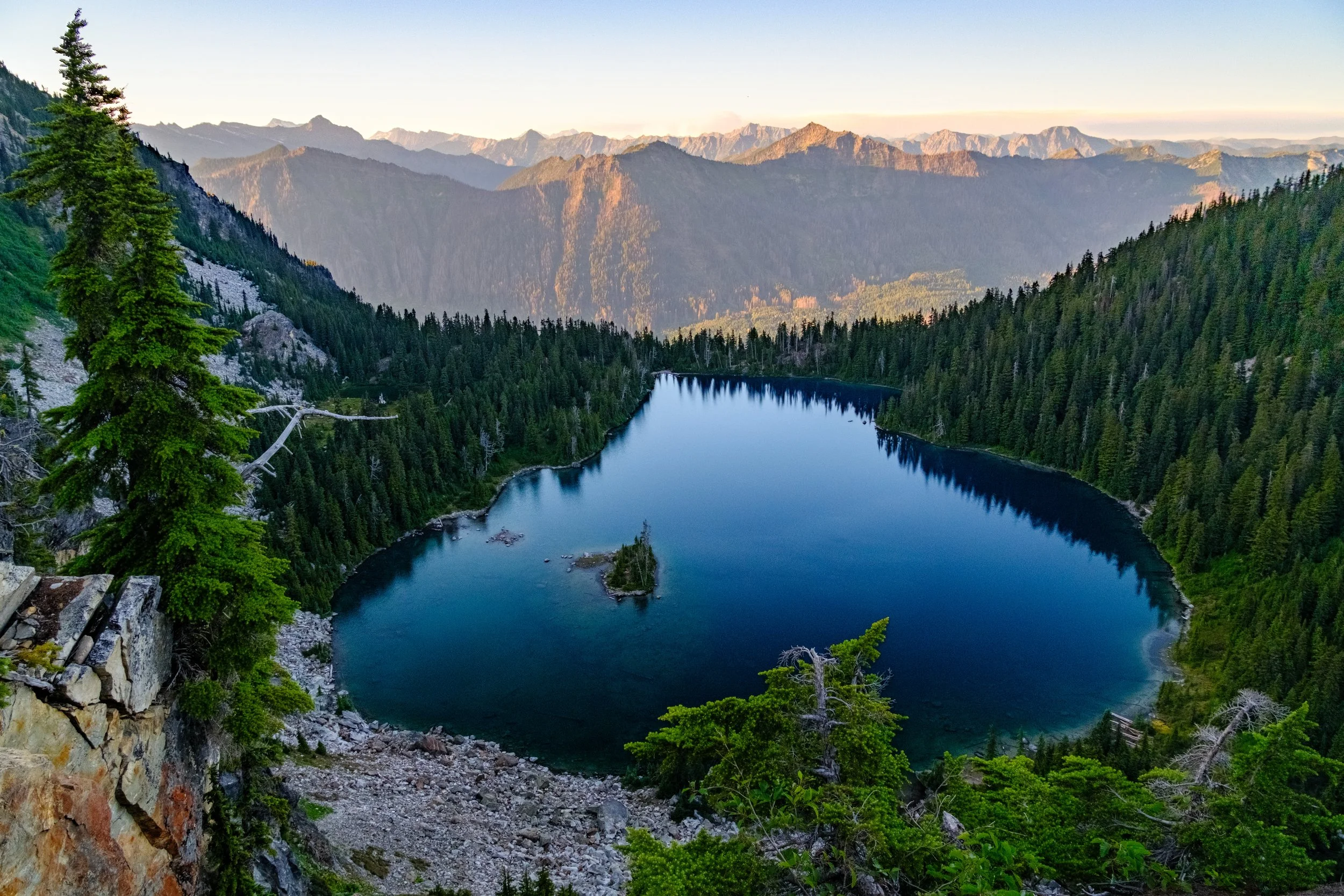 A mountain landscape featuring a large, calm lake surrounded by dense evergreen forests. Tall mountains are visible in the background under a clear sky.