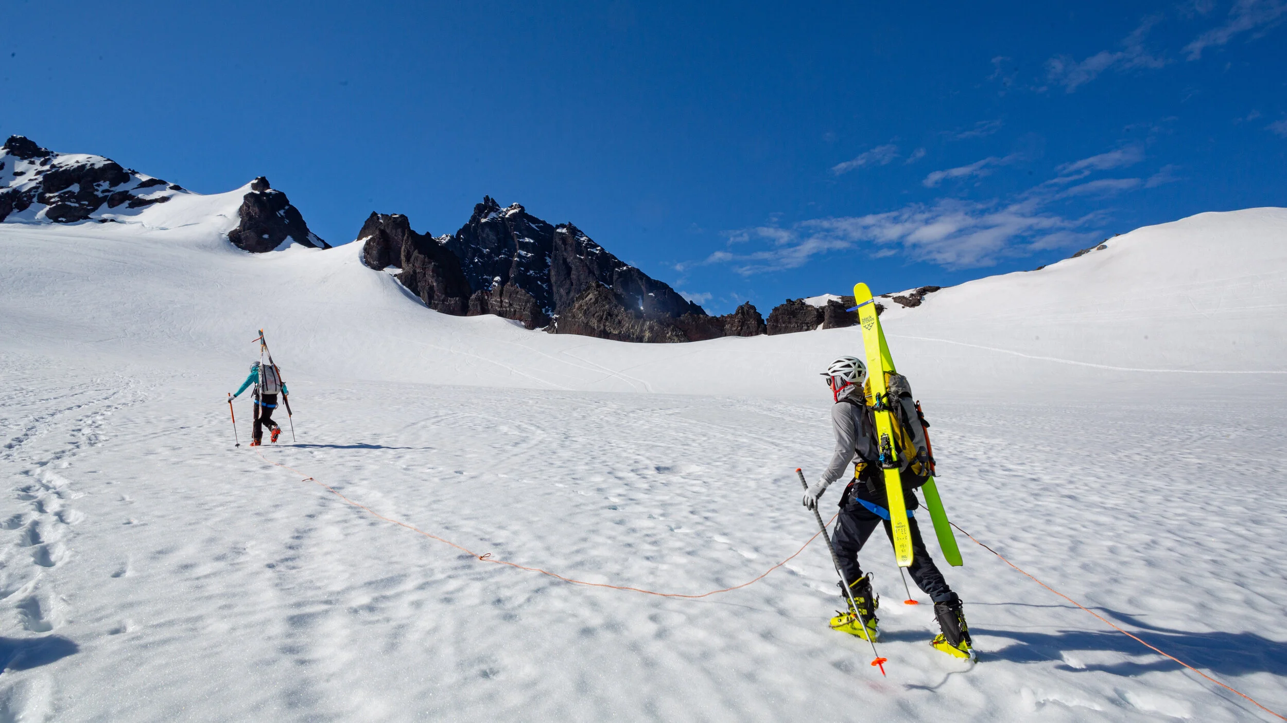 Two skiers in snow-covered mountainous terrain, one carrying skis on their back, ascending towards rugged peaks under a partly cloudy blue sky.