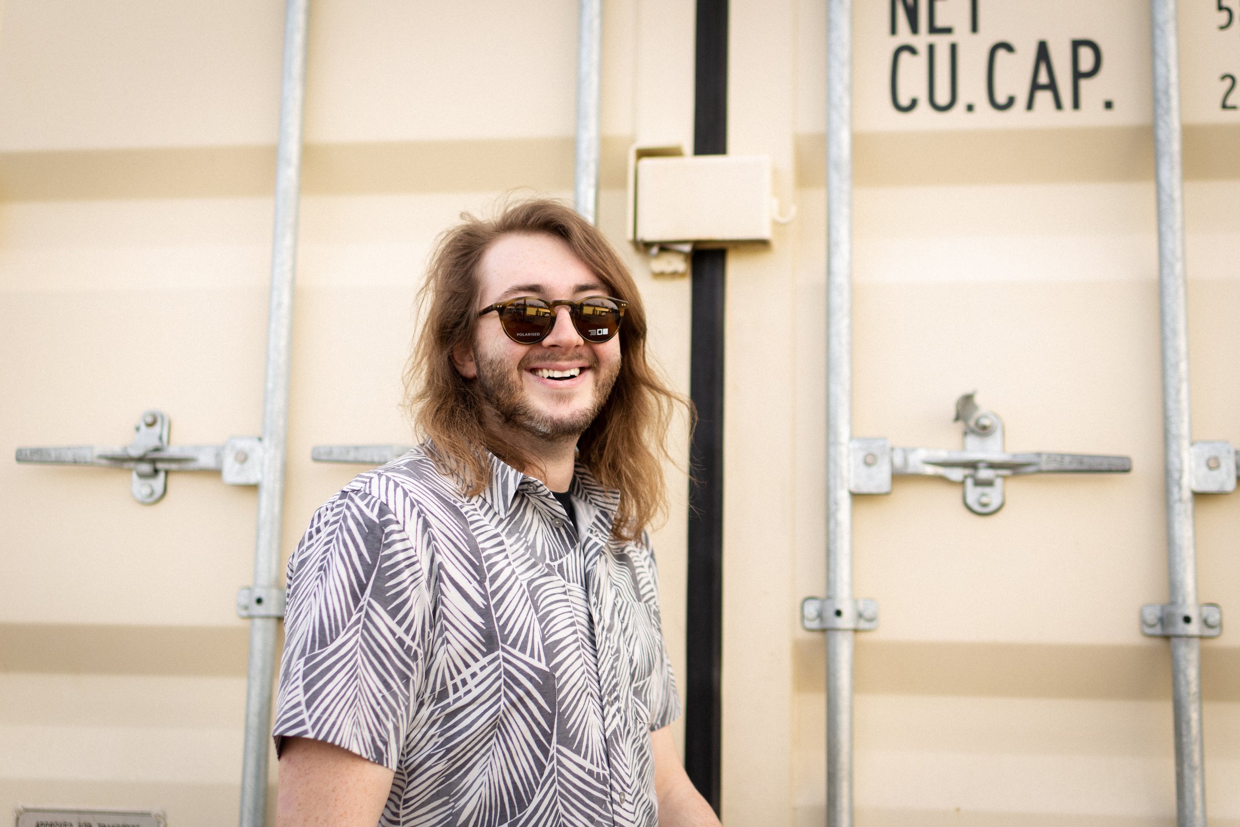 A smiling man with long hair, beard, wearing sunglasses and a patterned shirt, standing in front of a beige wall with metal bars and a sign.