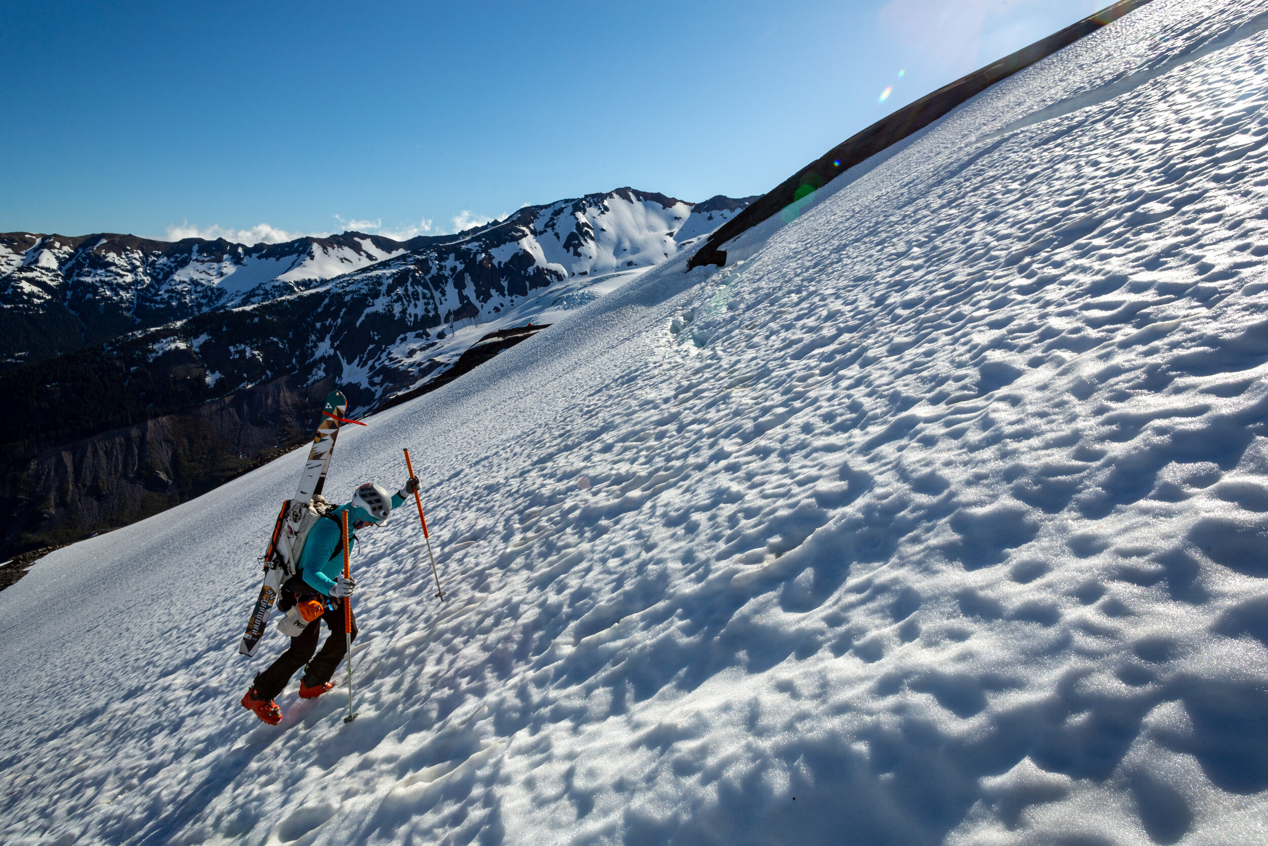 A mountain climber in a blue jacket, helmet, and gloves carrying skis, ascending a snowy slope with a mountainous landscape in the background.