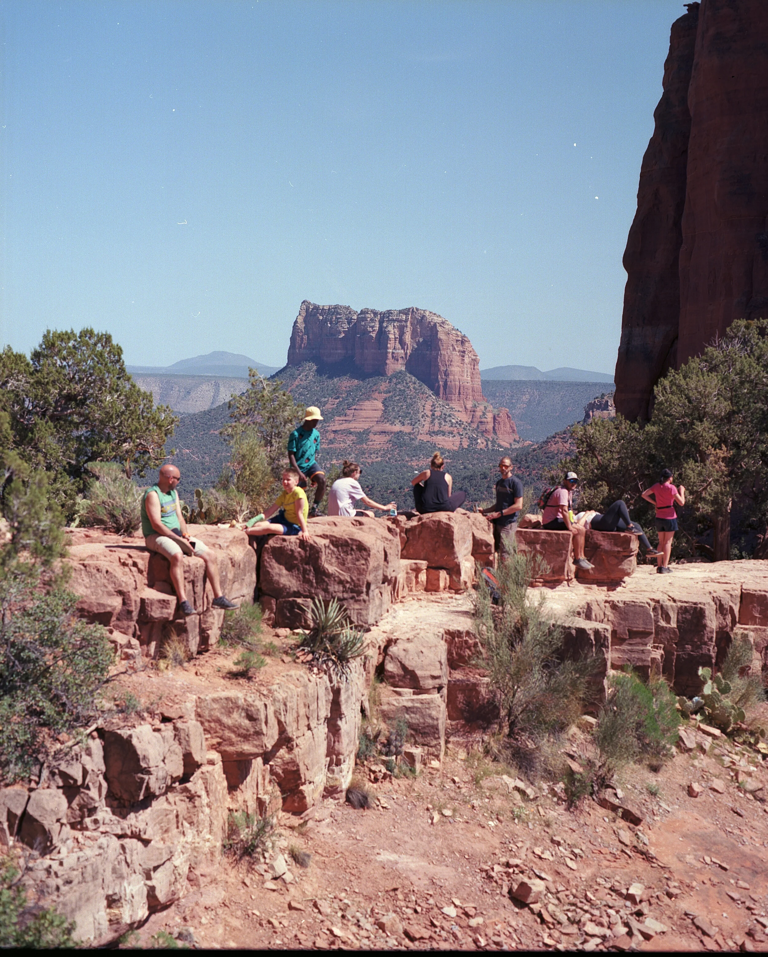 A group of tourists resting and relaxing on red rocks in front of a desert landscape with large rock formations and sparse vegetation.