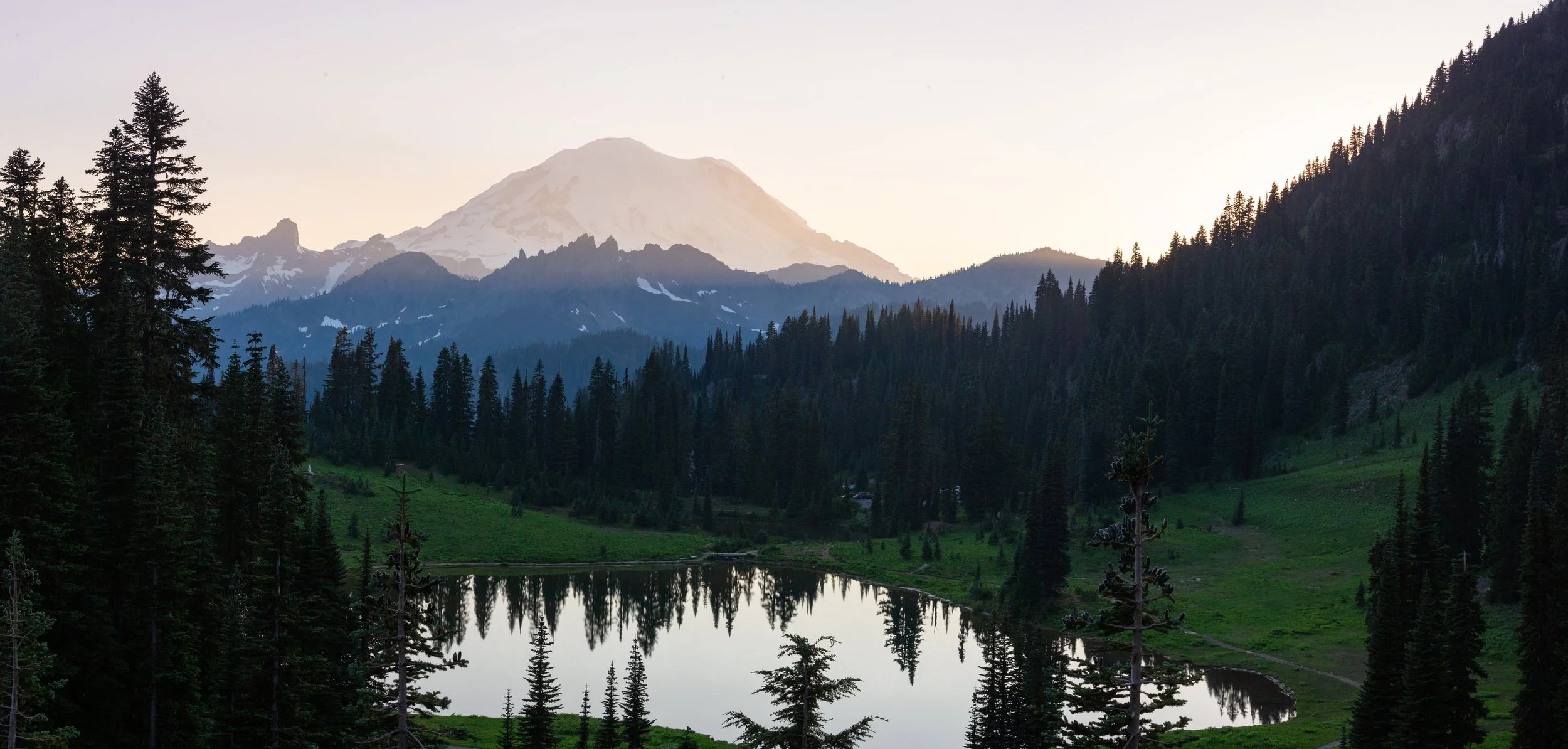Snow-capped mountains behind a dense forest and a small lake reflecting the trees, during sunset.