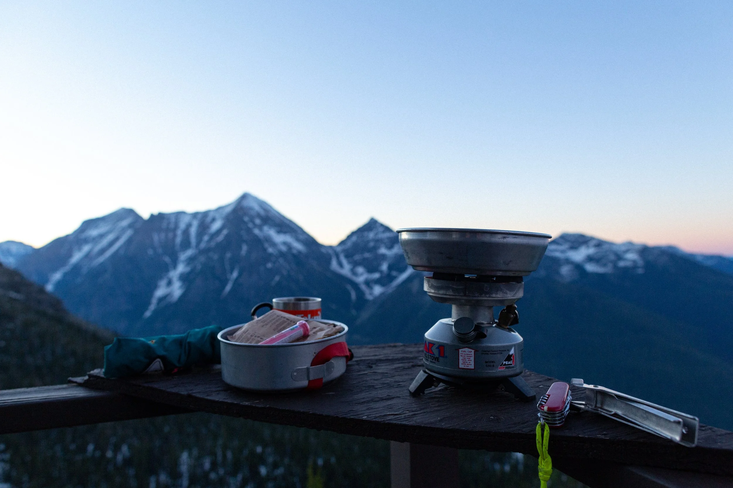 A camping stove, a mug, and supplies on a wooden surface with mountain peaks in the background at dusk.