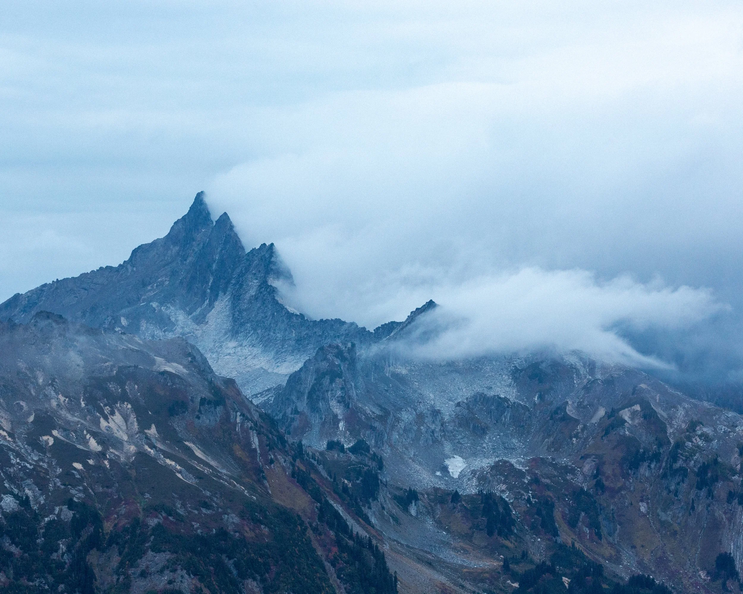 Snow-capped mountain peaks partially covered by clouds against a cloudy sky.