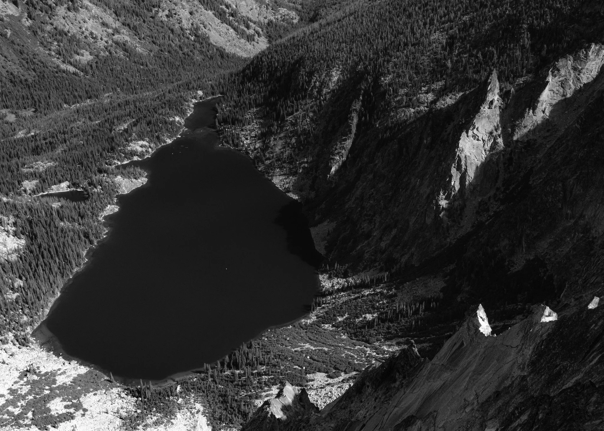 Aerial view of a lake surrounded by mountains and forested terrain, with steep cliffs and rugged landscape.