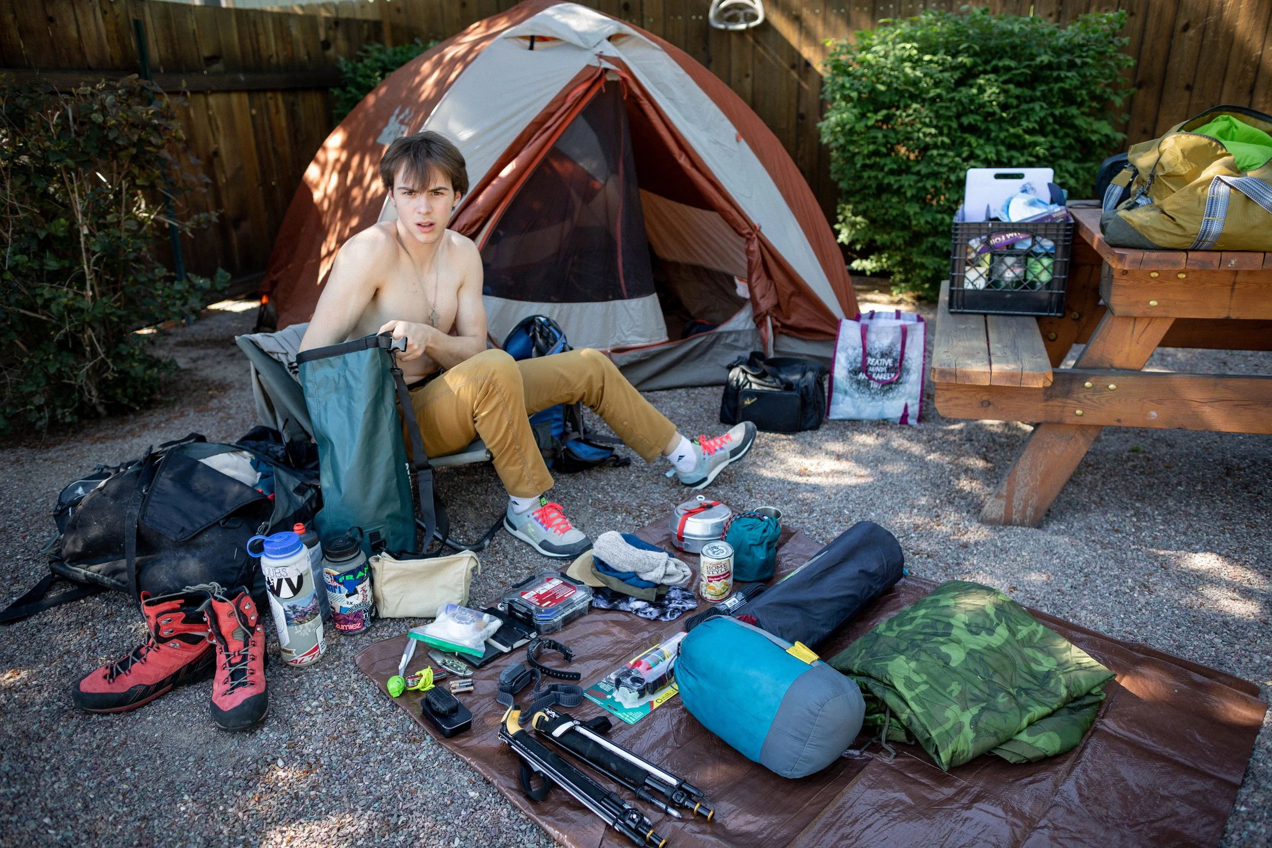 A young man with brown hair and light skin, sitting shirtless on a camping chair, surrounded by camping gear. There's a tent, various bags, shoes, and outdoor equipment laid out on the ground and table. Glacier National Park