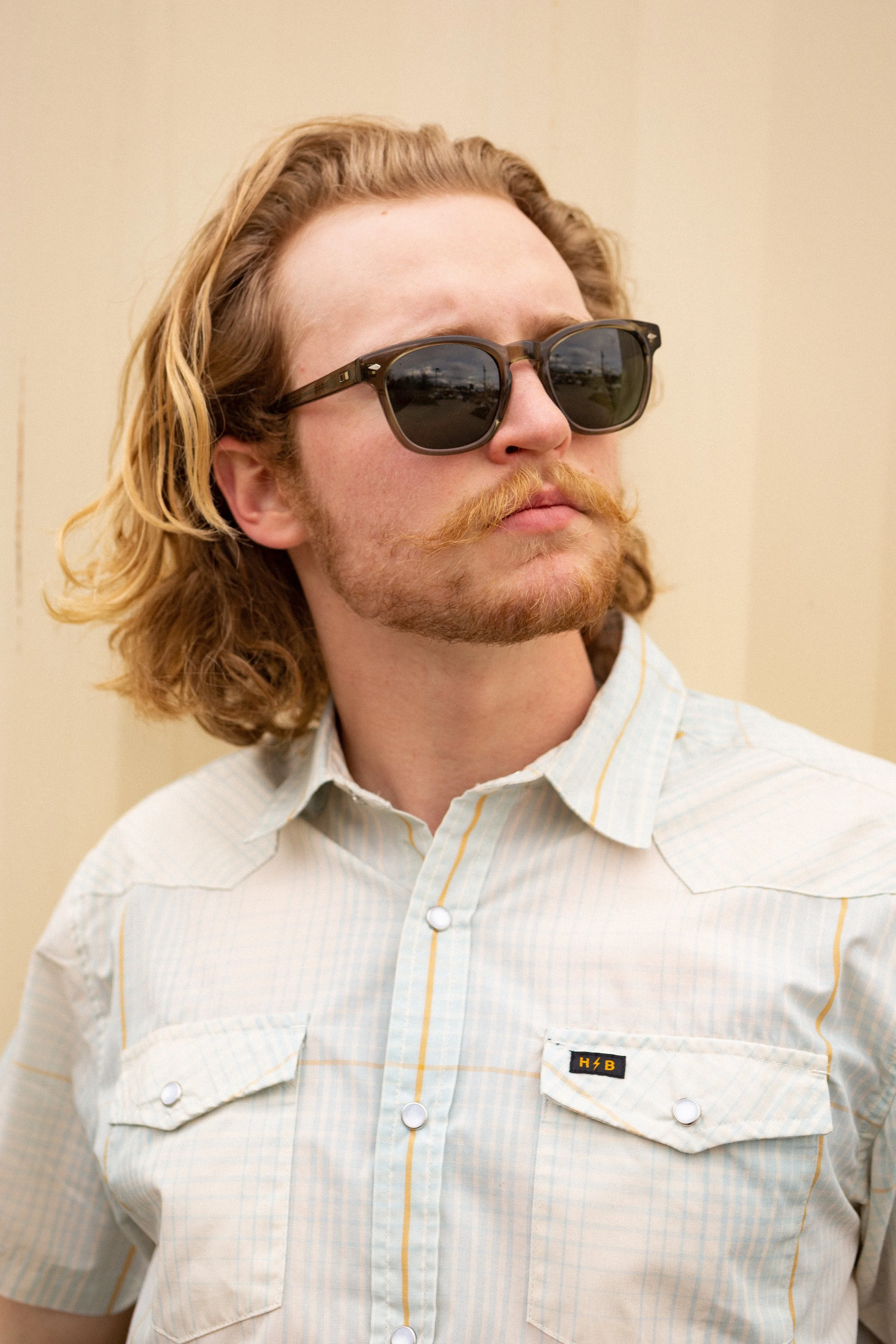 A man with wavy, shoulder-length hair, a reddish beard and mustache, wearing sunglasses and a cream-colored, short-sleeved, button-up shirt with a small black and yellow patch on the pocket.