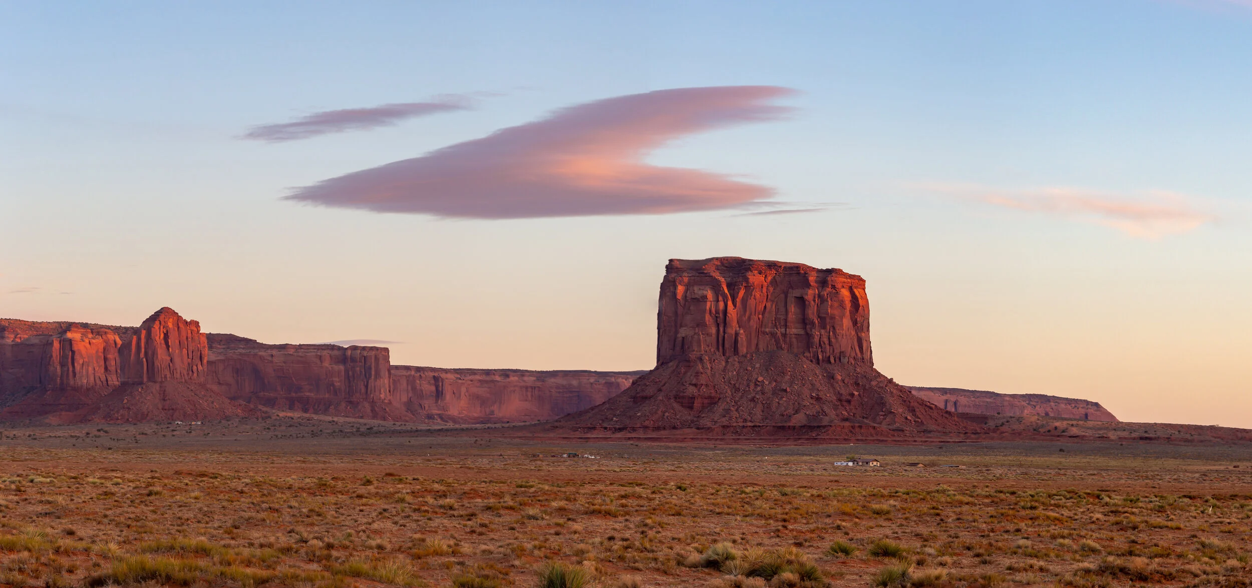 Desert landscape with large red rock formations and a flat plain in the foreground, under a partly cloudy sky at sunset or sunrise