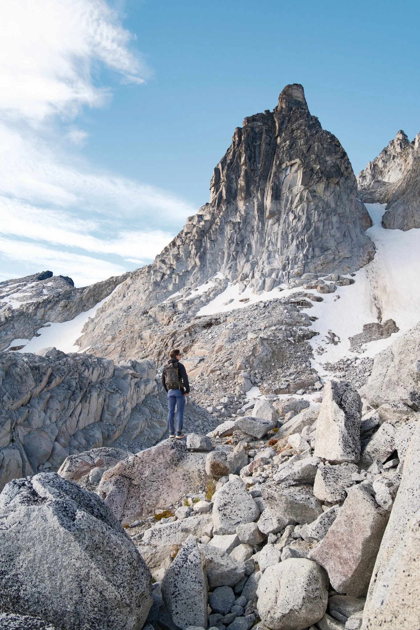 A hiker standing among large rocks in a mountainous landscape with snow patches and a large rocky peak in the background under a blue sky with scattered clouds.