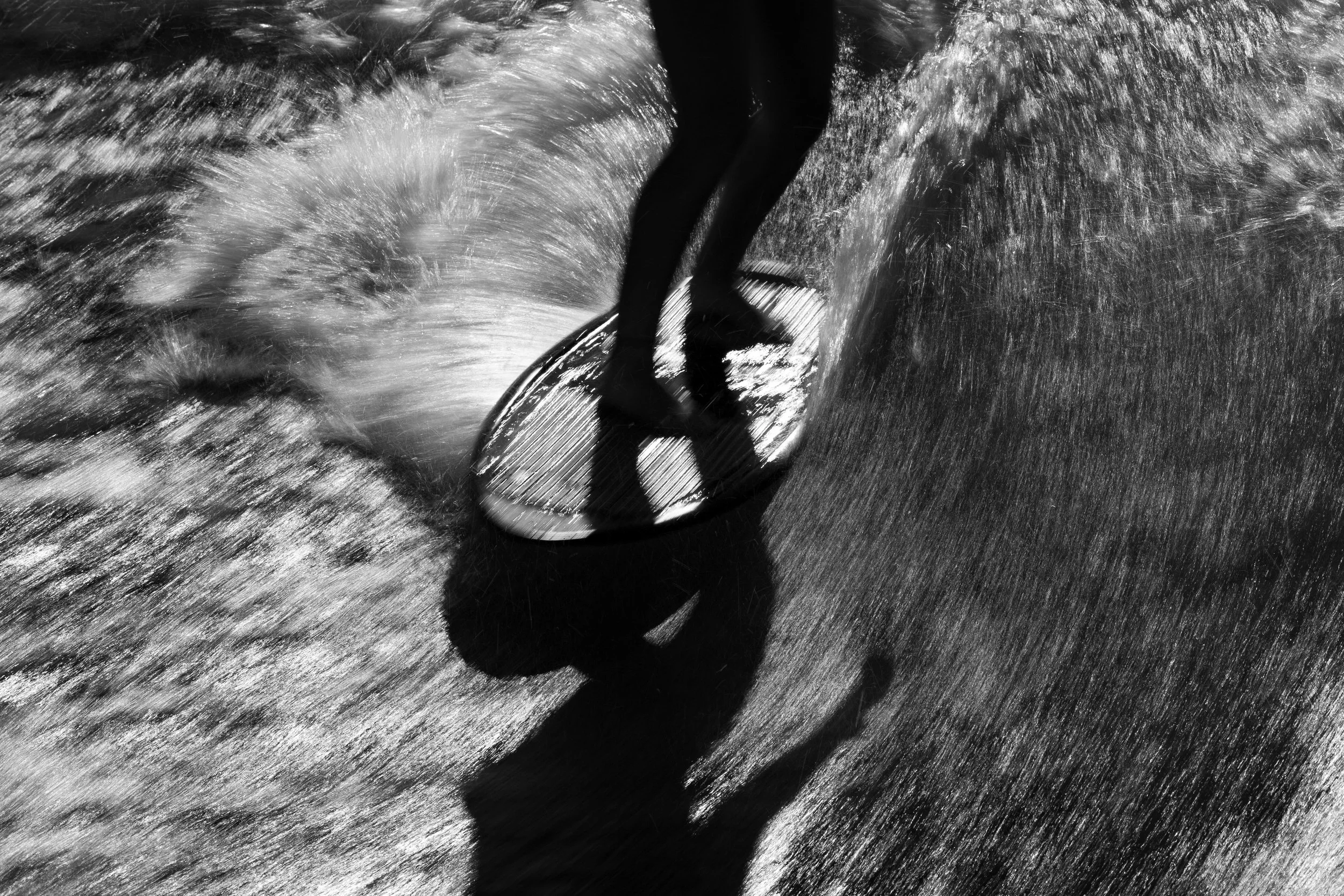 A person surfing on a wave, captured in black and white with the sun casting a shadow of the surfer on the sand.