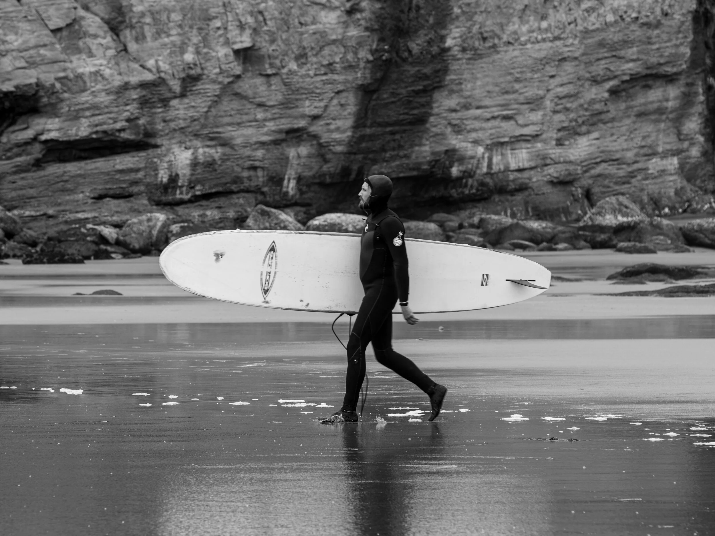 A person in wetsuit walking on the beach carrying a surfboard under their arm with a rocky cliff in the background.