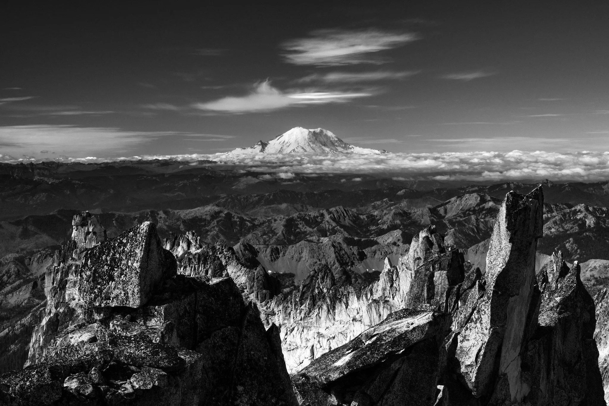 Black and white landscape photo of rugged mountain peaks with a snow-capped mountain in the background, Mount Rainier. 