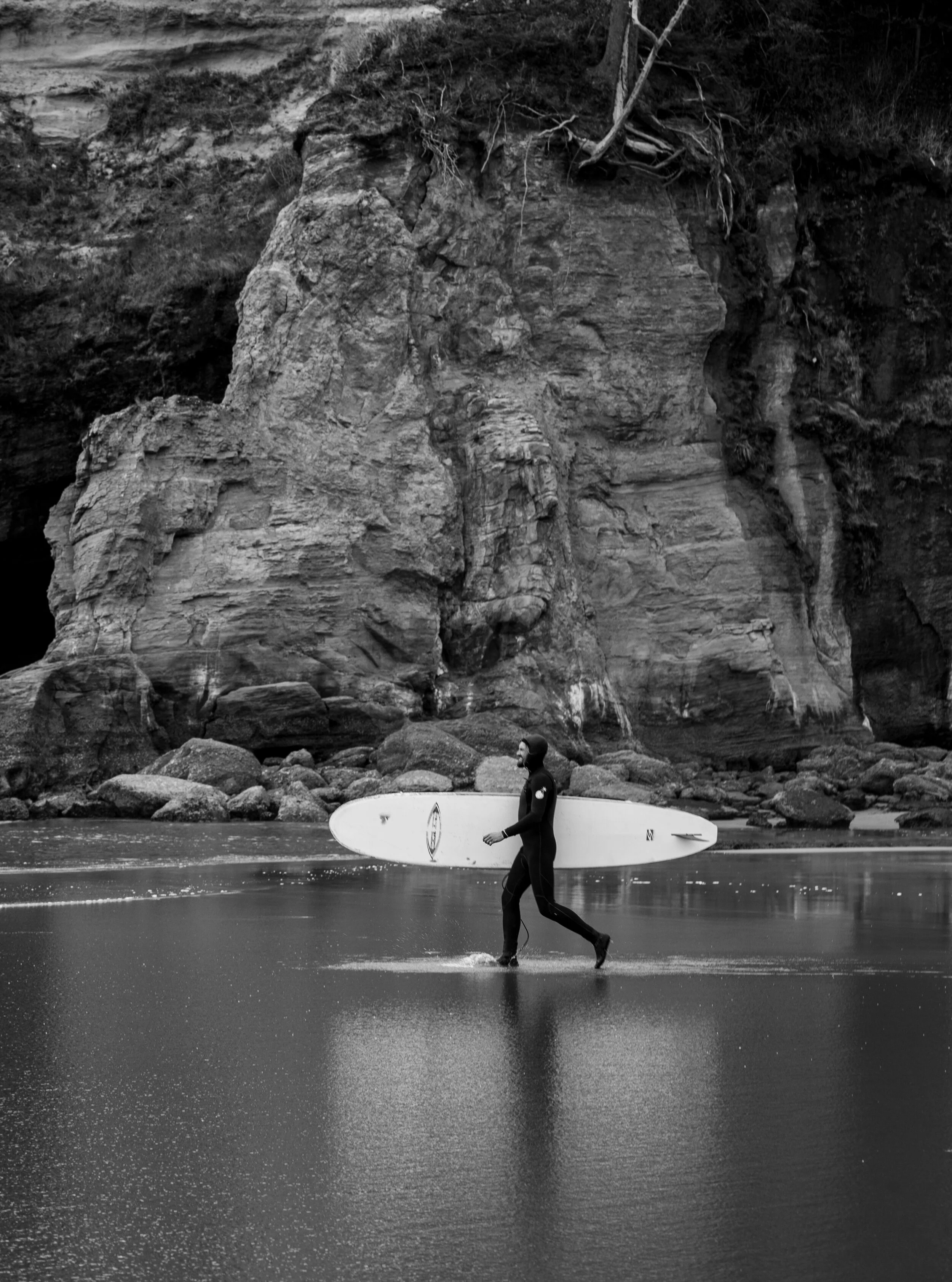 A person in a wetsuit is walking through shallow water carrying a surfboard under an overhanging rocky cliff on the Oregon Coast. Otter Rock area
