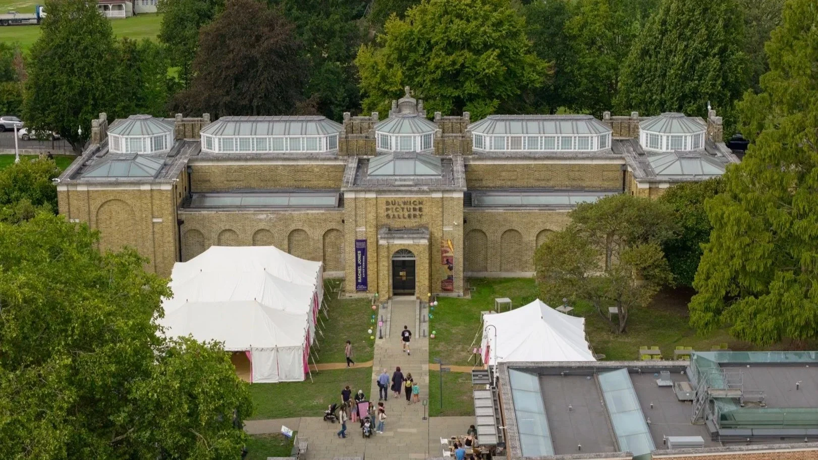 Aerial view of Dulwich Picture Gallery, a historic building with glass-roofed extensions, surrounded by green trees and outdoor tents, with visitors walking on the pathways.