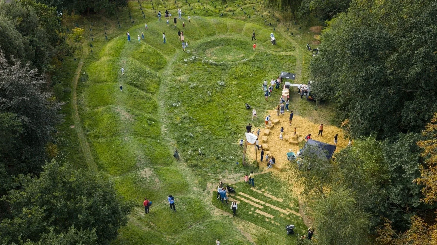 An aerial view of a park or festival area with people walking around, playing, and sitting on hay bales. The area has grassy and dirt sections, surrounded by trees, and some equipment and vehicles visible.