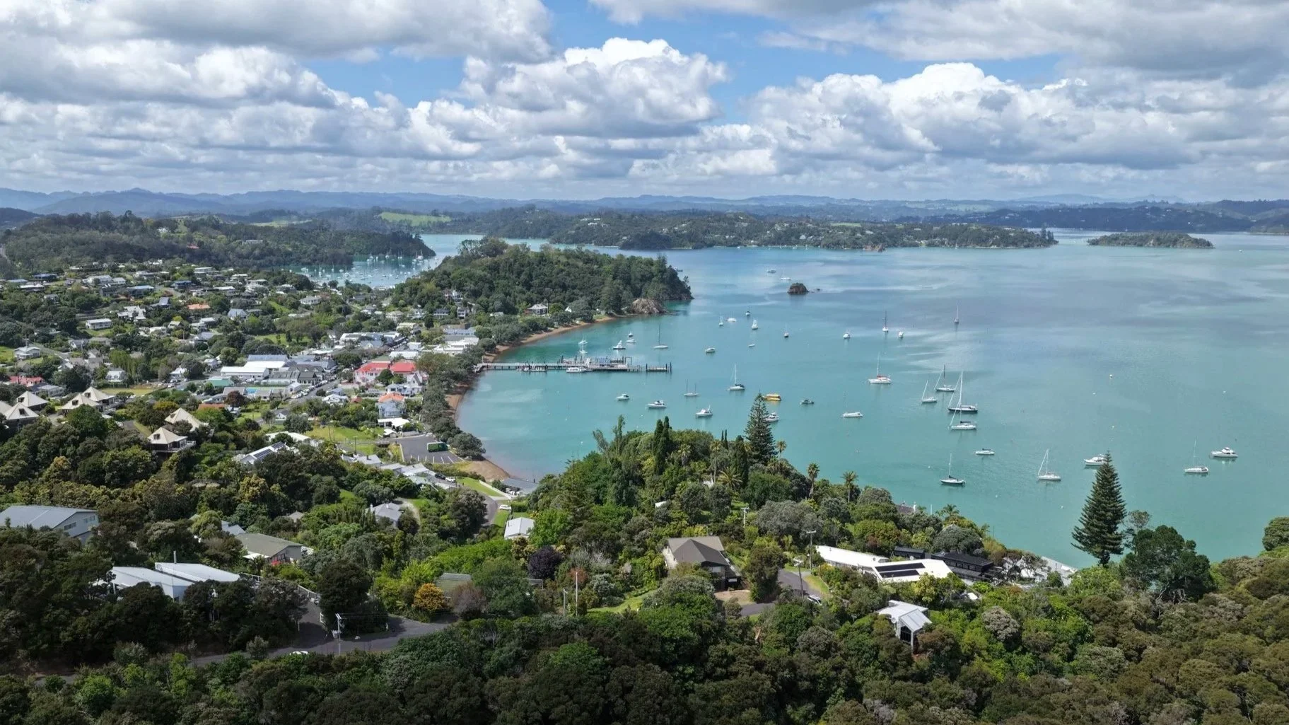 Aerial view of a coastal town with houses, trees, and a marina with sailboats on a calm bay, under a partly cloudy sky.