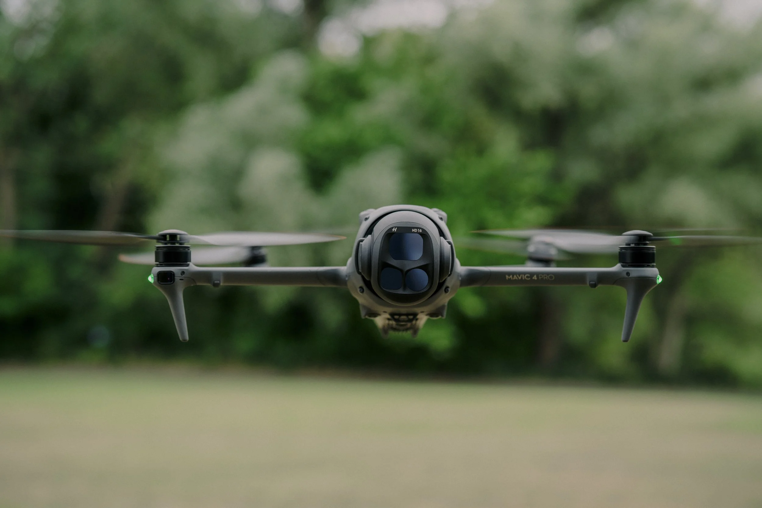 A drone flying in the air with a blurred green background of trees and grass.