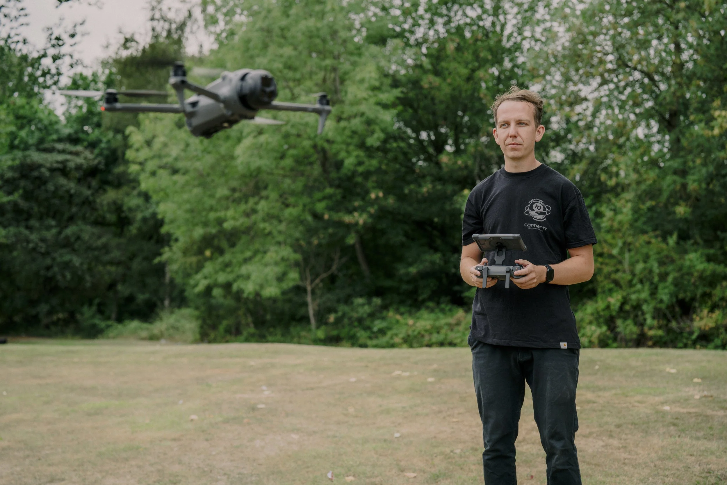 A man operating a drone outdoors in a park or forested area, holding a remote control while the drone flies nearby.
