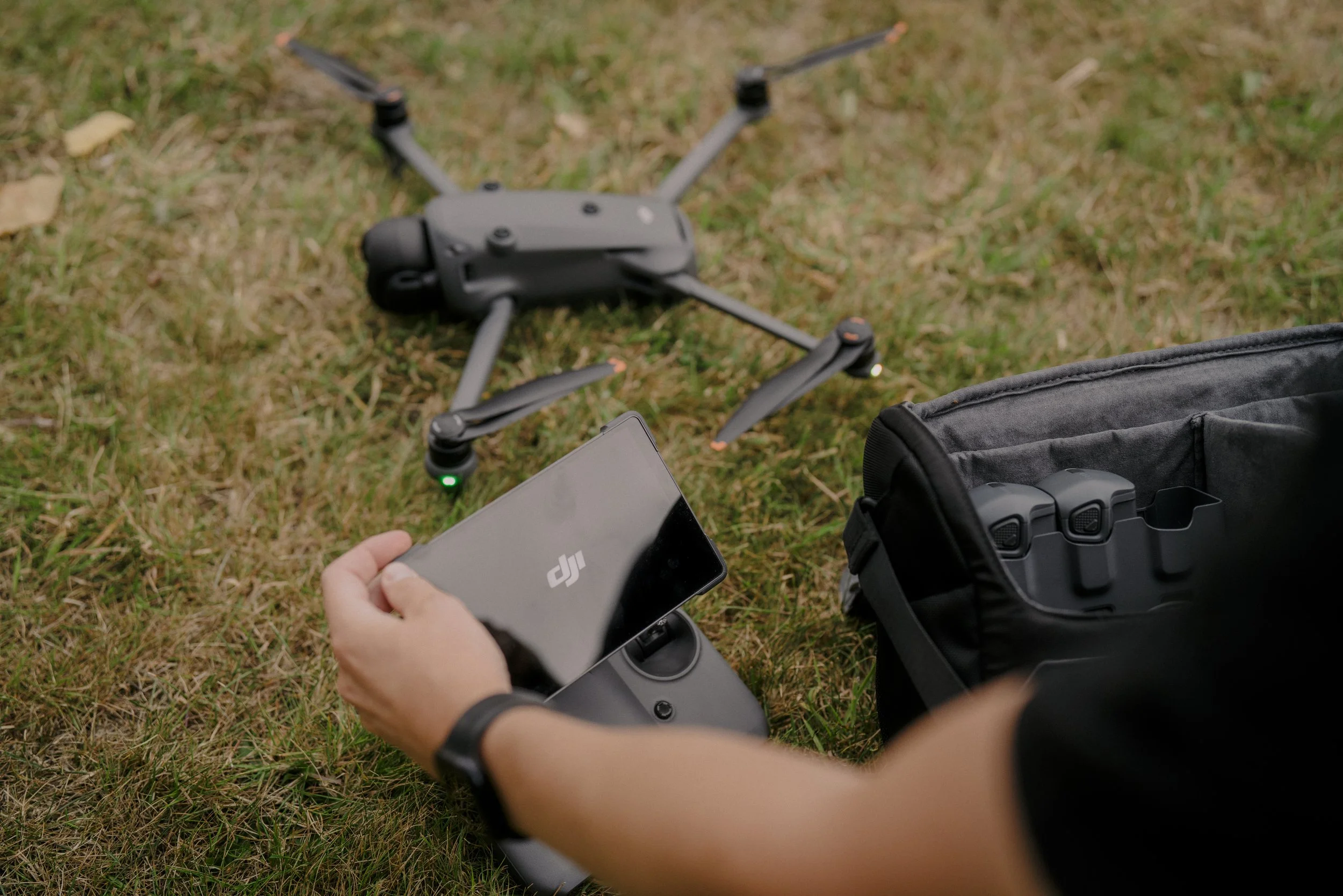 A person is preparing a DJI drone outside on grass, holding a remote controller with a smartphone attached, with the drone on the ground nearby and a carrying case open beside them.