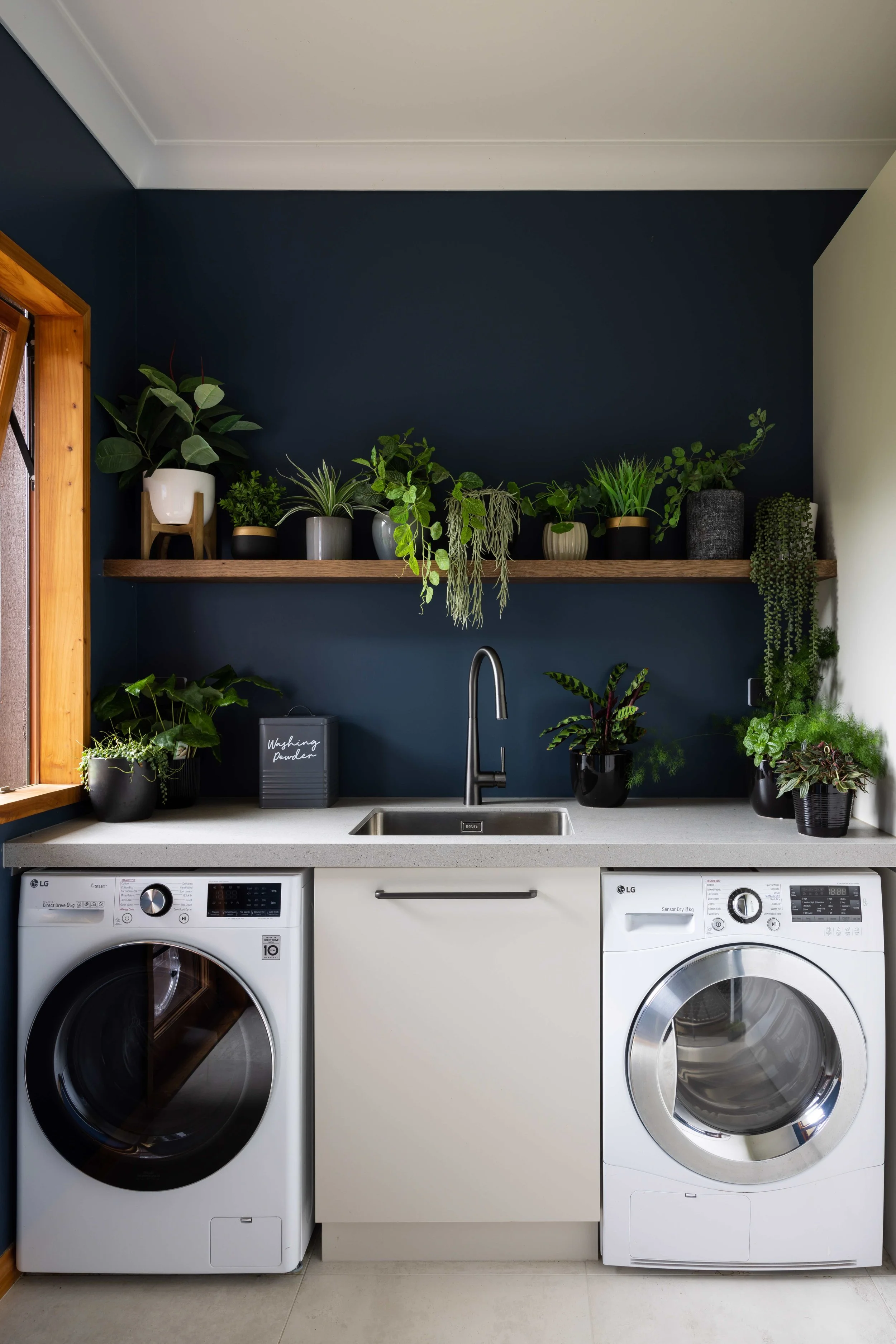 Laundry room with dark blue wall, wooden window frame, and white LG washer and dryer, white countertop, black faucet, and various potted plants on shelf and countertop.