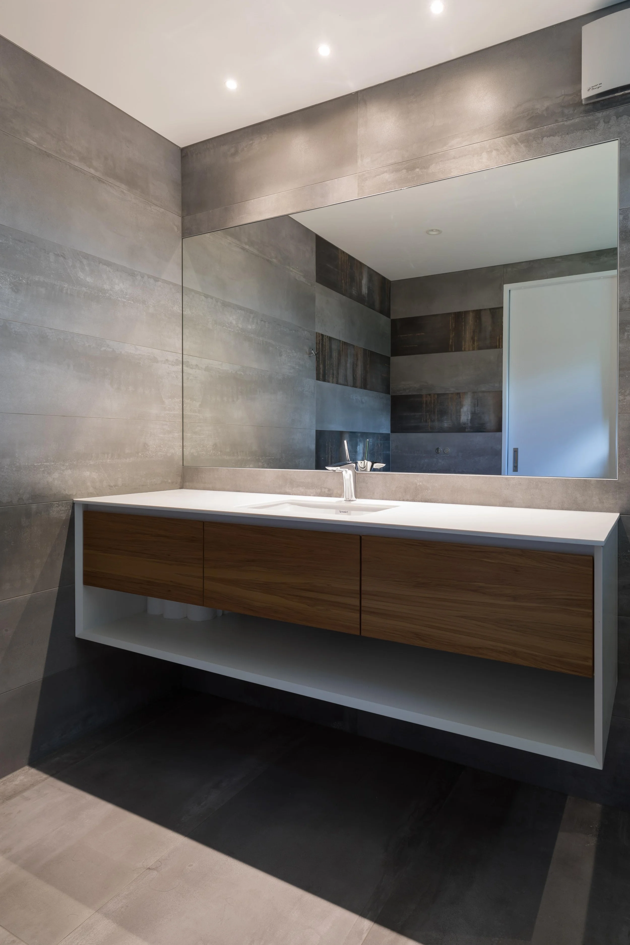Modern bathroom with a large mirror, grey tiled walls, a white countertop with a wooden cabinet underneath, and a faucet and sink.