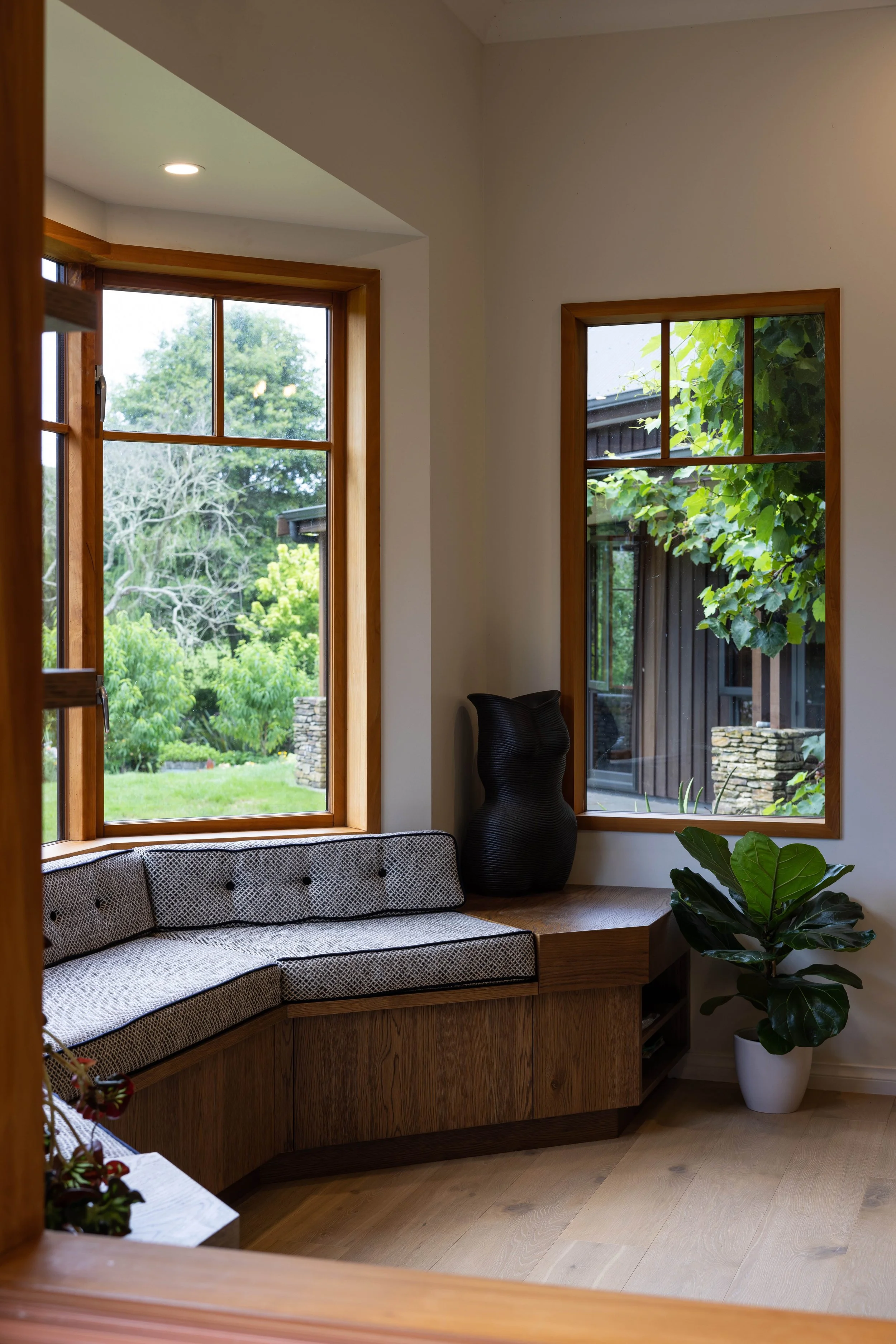 A cozy corner with built-in seating by two large windows showing greenery outside, decorated with a black vase, a potted plant, and wooden furnishings.