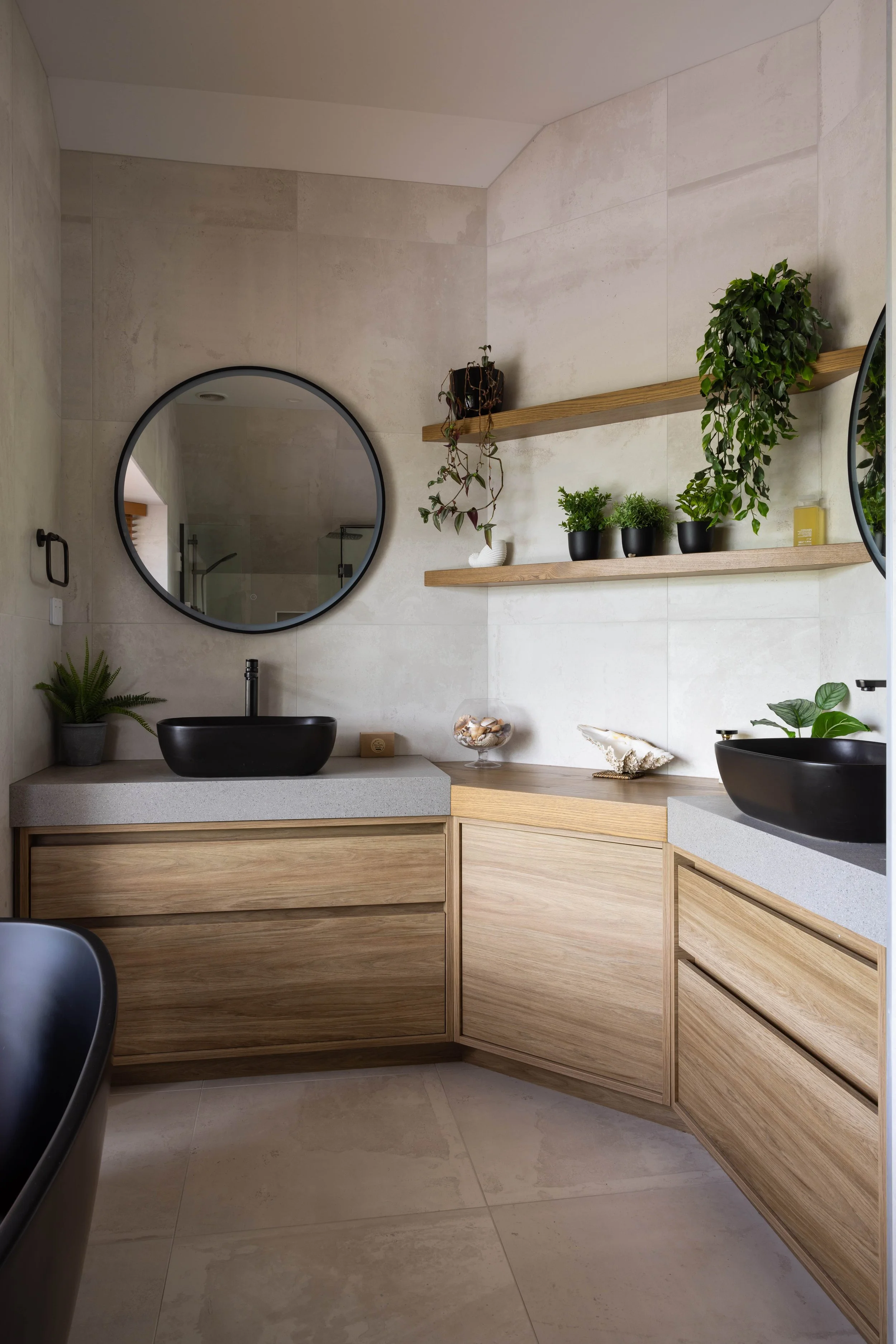 Modern bathroom with beige tiled walls, wooden cabinetry, black vessel sinks, round mirror, and potted green plants on wooden shelves.