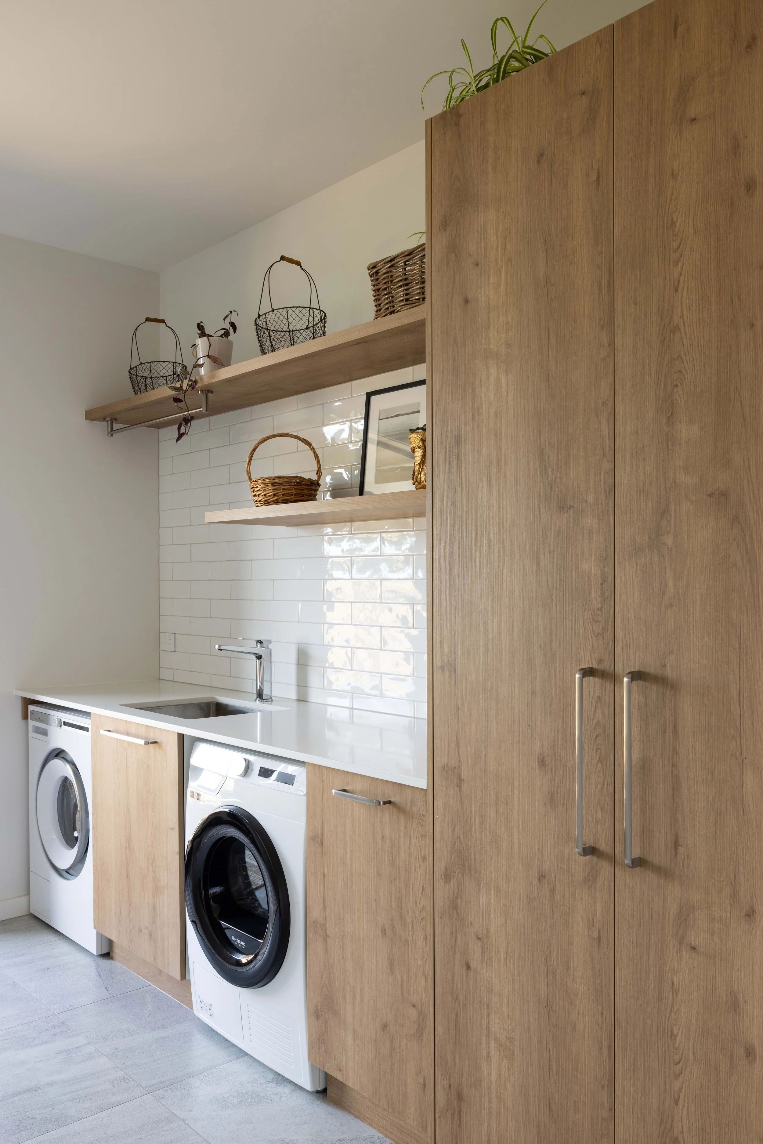 A laundry room with wooden cabinets, two open shelves with decor, a white tiled backsplash, a sink, and a washing machine and dryer.