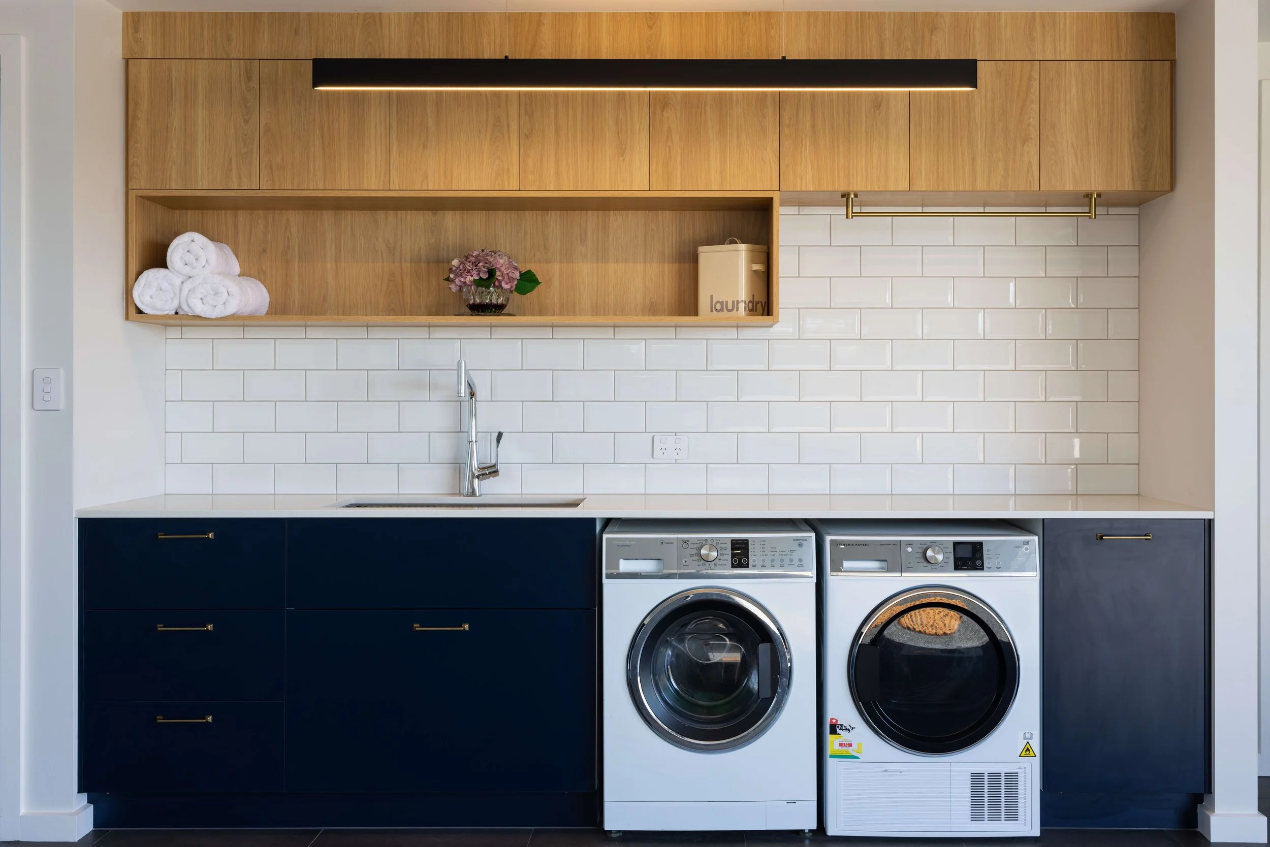 Laundry room with dark blue cabinets, white countertop, front-loading washer and dryer, white subway tile backsplash, open wooden shelves, towels, a flower arrangement, and a laundry bin.